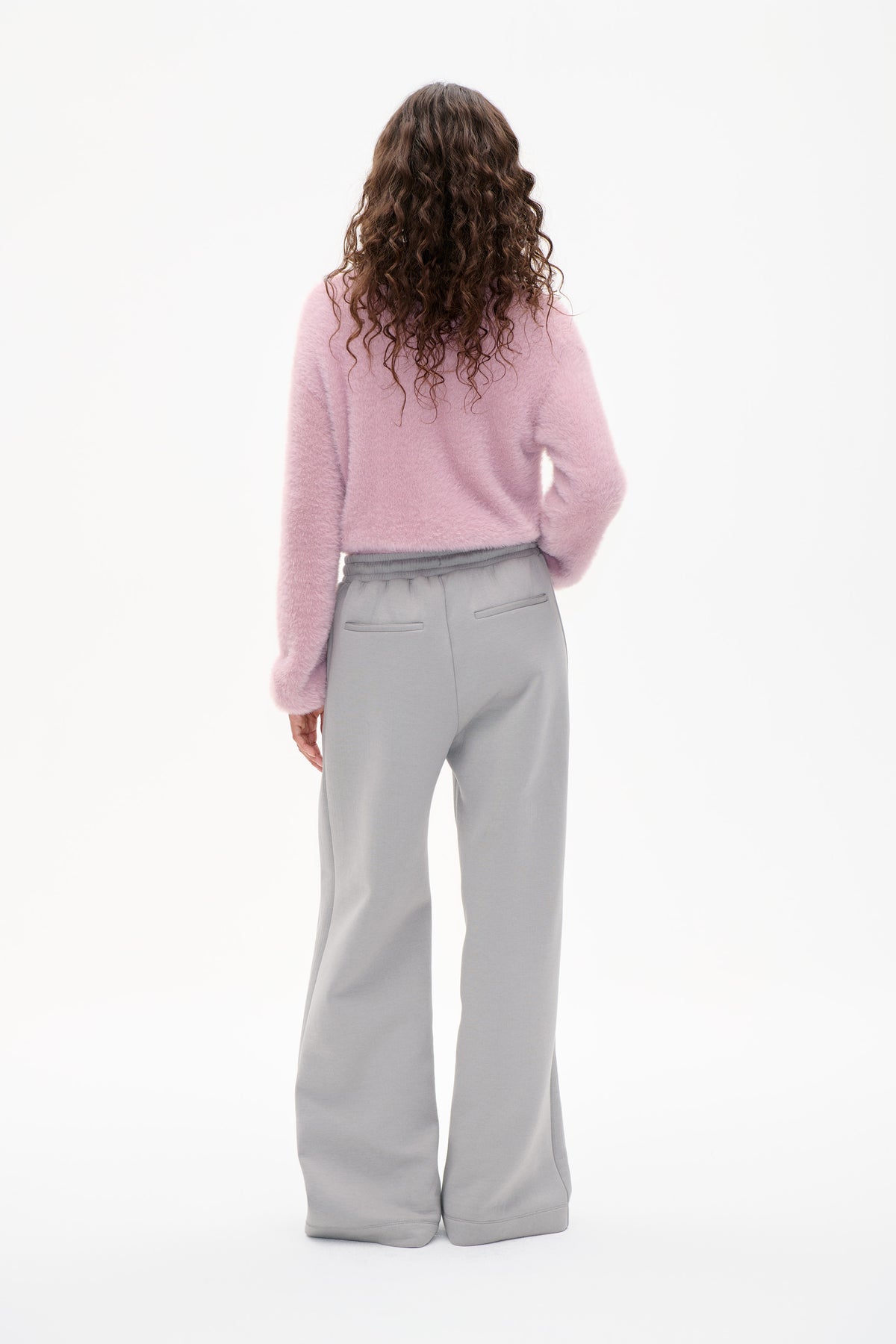 Rückenansicht eines Models, das einen flauschigen hellrosa Pullover und eine hellgraue, weit geschnittene Hose mit elastischem Bund und Leistentaschen trägt.