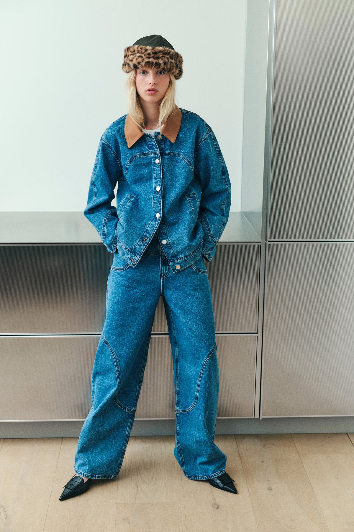 Model trägt eine blaue Denimjacke mit hellbraunem Kragen, dazu passende Jeans mit weitem Bein und einen dunklen Hut mit leopardengemusterter Krempe.