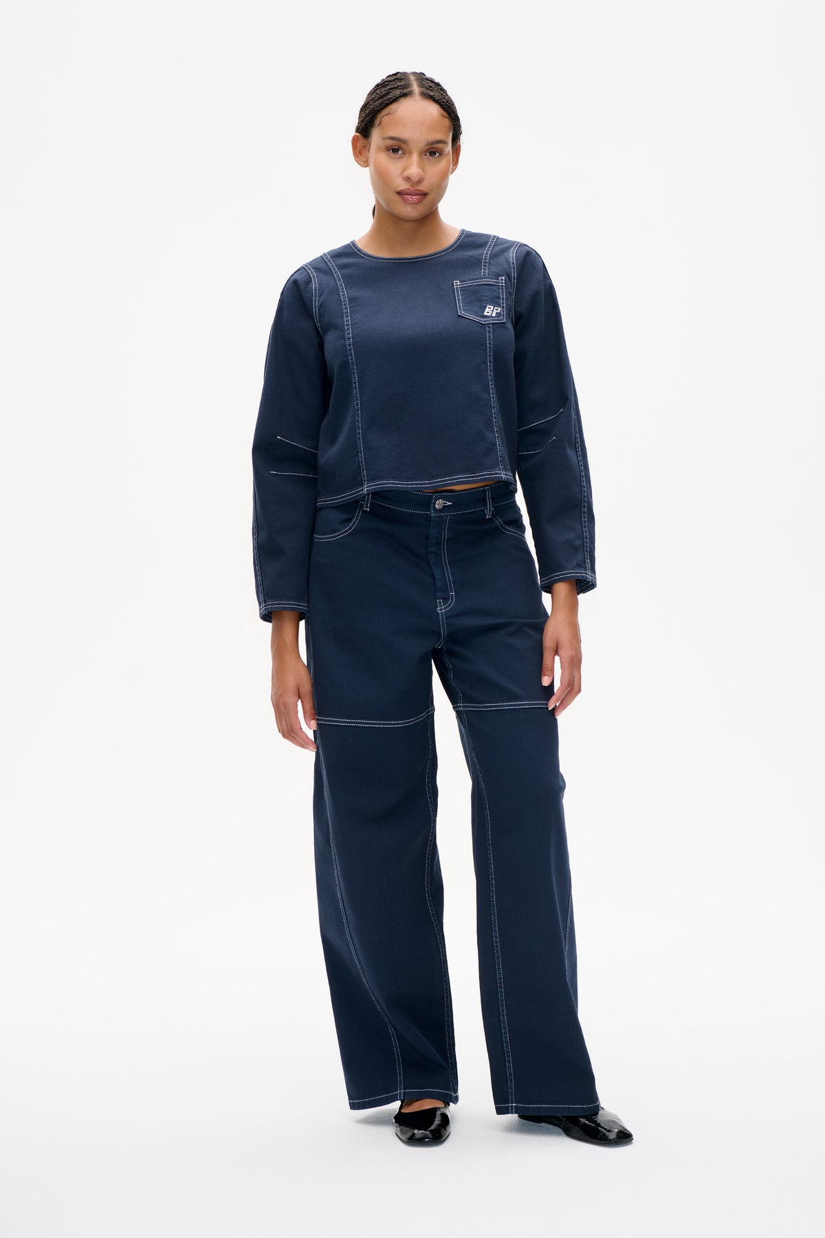 Vorderansicht eines Models, das ein marineblaues Langarmtop mit Kontrastnähten und Brusttasche trägt, kombiniert mit einer weit geschnittenen Hose.
