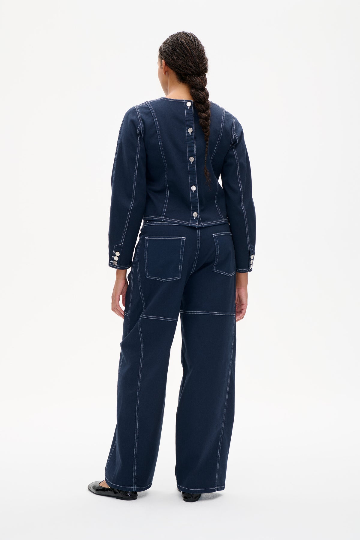 Back view of a dark indigo denim jacket and matching wide-leg trousers with contrast white stitching and silver-tone buttons.