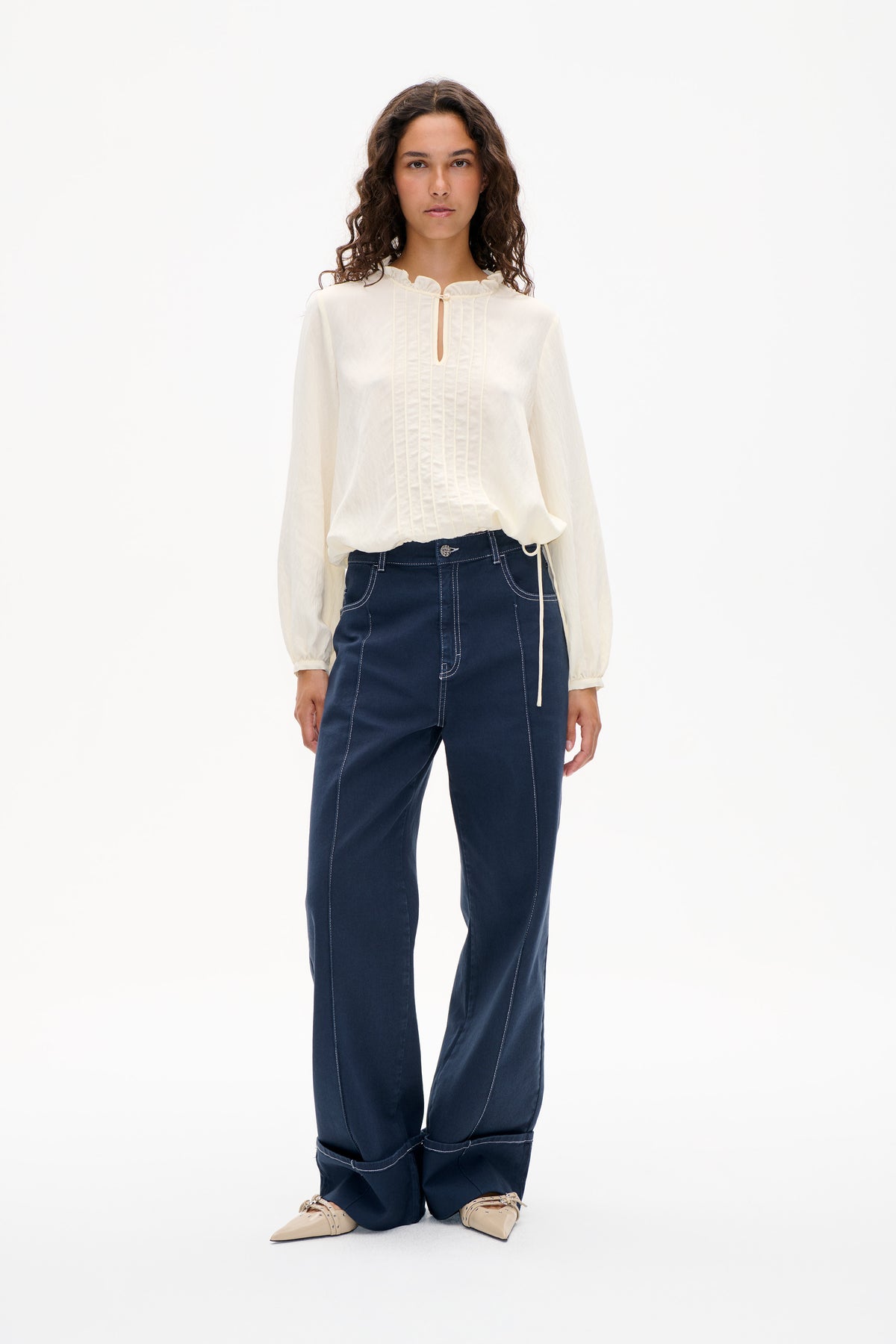 Model wearing an ivory long-sleeved blouse with a ruffled neckline and pintuck front, paired with dark indigo wide-leg jeans with contrast stitching.