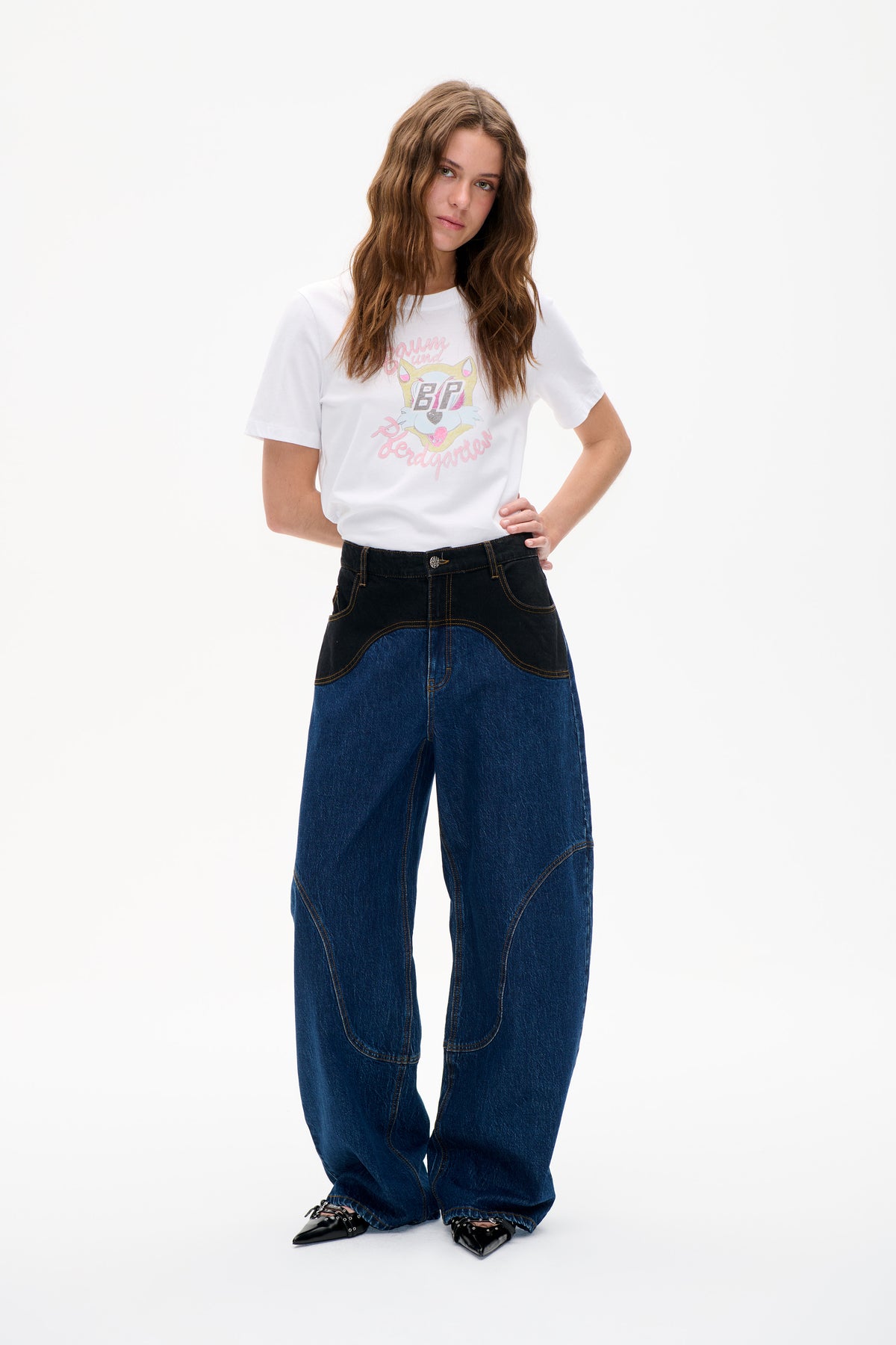 Model trägt ein weißes kurzärmeliges T-Shirt mit pastellfarbenem Katzenmotiv und Schriftzug, kombiniert mit einer weiten blauen Jeans mit schwarzen Kontrastpaneelen.