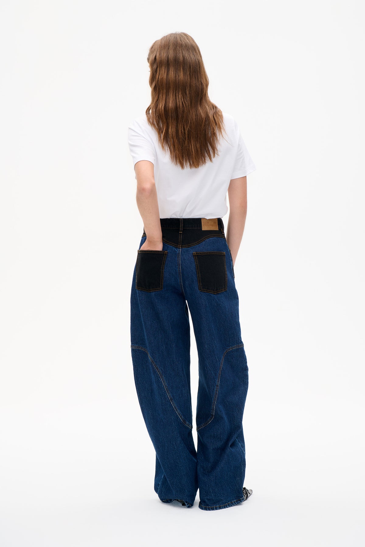 Rückenansicht eines Models, das ein weißes kurzärmeliges T‑Shirt und weit geschnittene blaue Jeans mit schwarzen Kontrast‑Einsätzen und aufgesetzten Taschen trägt.