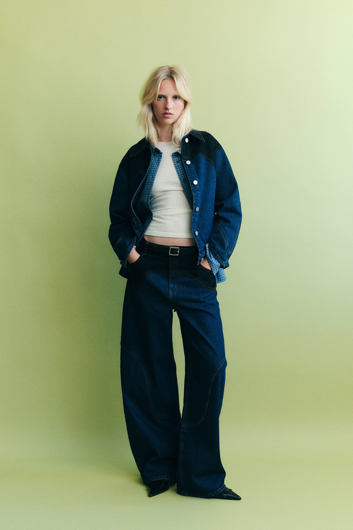 Das Model trägt eine zweifarbige Denimjacke in Navy und Blau über einem blau karierten Hemd und einem weißen Top, dazu Jeans mit weitem Bein vor einem blassgrünen Hintergrund.