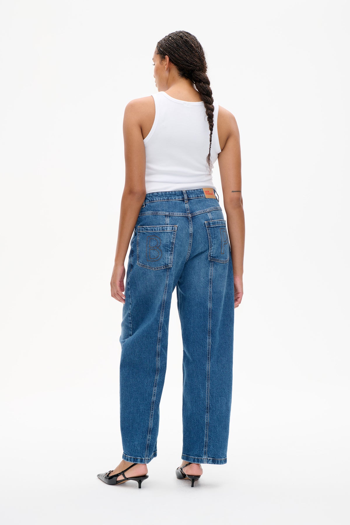 Back view of blue wide-leg jeans with oversized lettered patch pockets, styled with a white ribbed tank top.