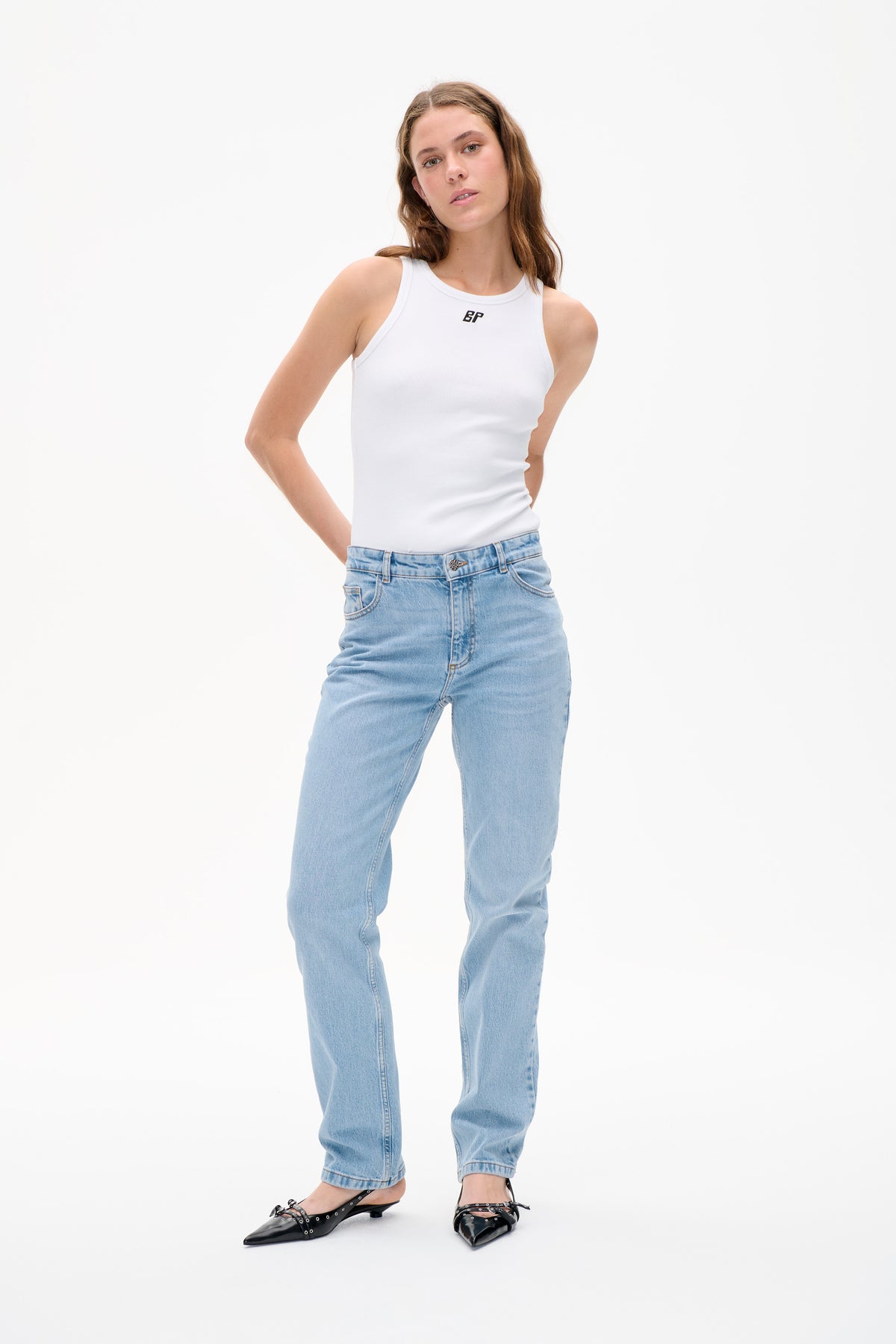 Model trägt ein weißes, geripptes ärmelloses Top mit kleinem schwarzem BP-Logo, hellblaue Jeans und schwarze spitze Ballerinas vor weißem Hintergrund.