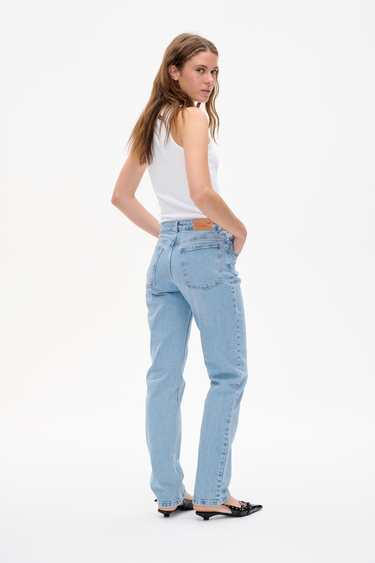 Model trägt ein weißes, geripptes Tanktop und hellgewaschene Jeans, von hinten gezeigt mit aufgesetzten Taschen und einem Etikett am Bund.
