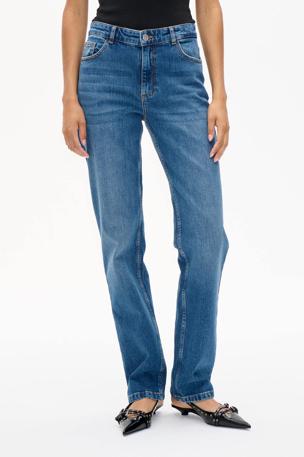 Voorpanddetail van blauwe denimjeans met vervaagde slijtagelijnen, contrasterende stiksels, riemlussen en een metalen merkknoop, gecombineerd met een zwart topje.
