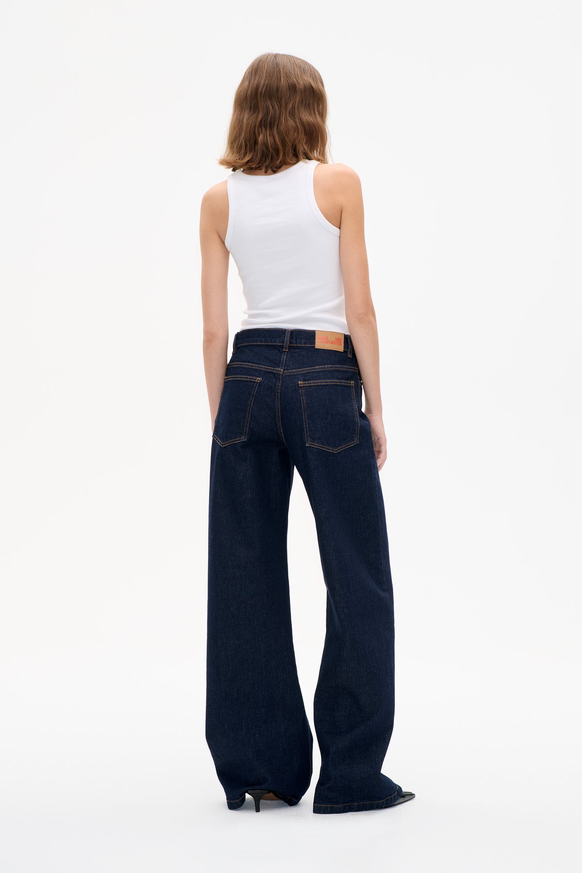 Rear view of a model in dark indigo wide-leg jeans with contrast stitching and patch pockets, paired with a white ribbed tank top.