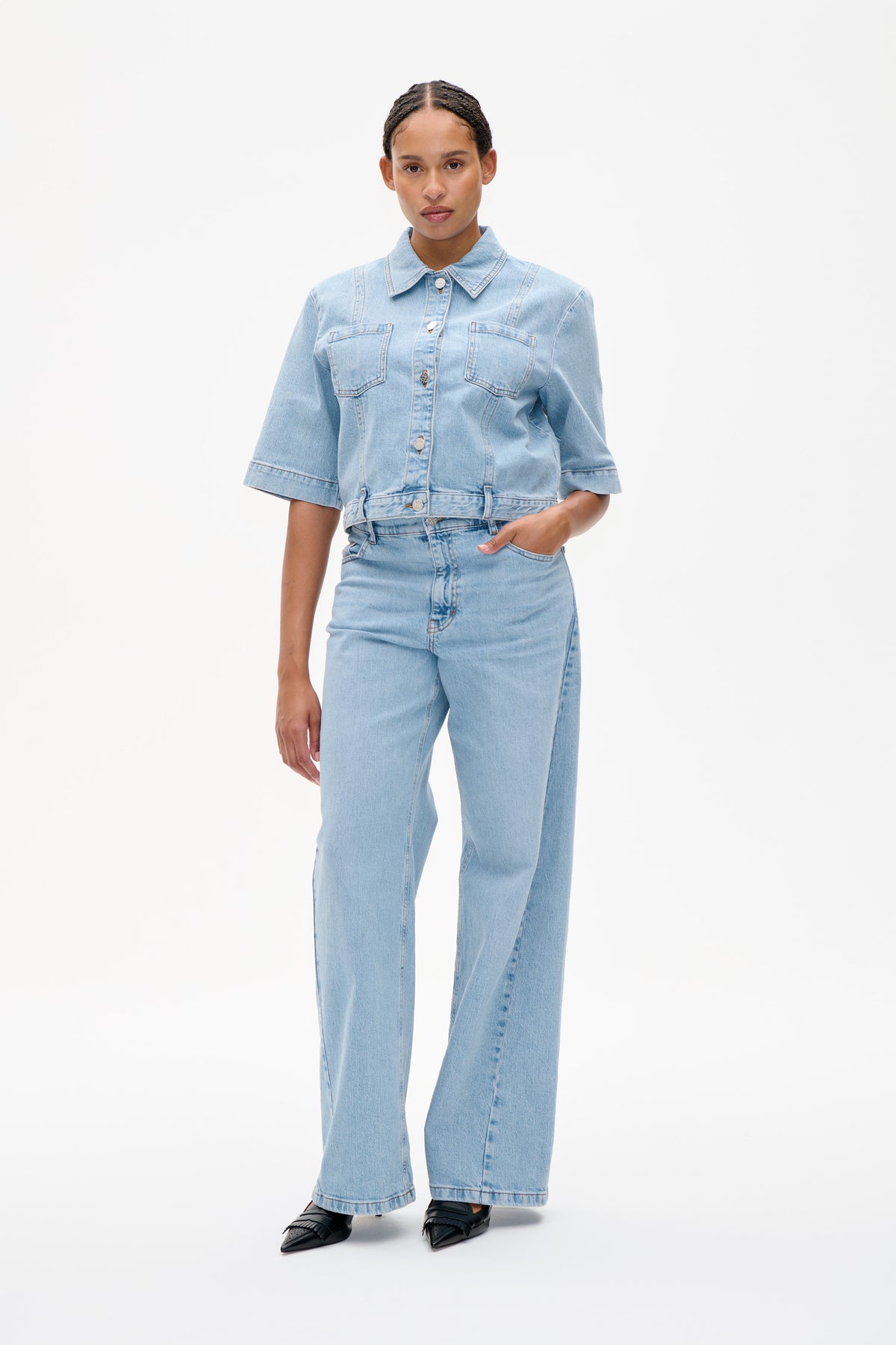 Front view of a model wearing a light blue denim jacket with short sleeves, a pointed collar, button closure and two chest pockets, with matching wide-leg jeans.