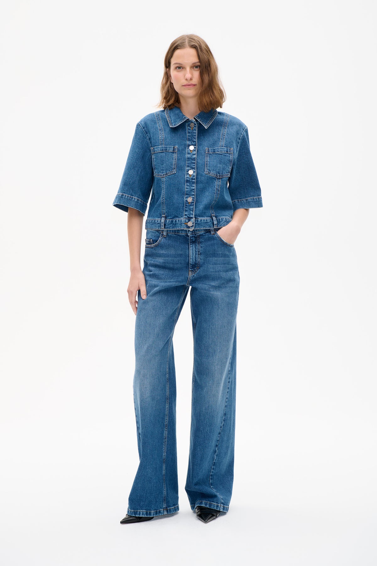 Model wearing a blue denim button-front jacket with a pointed collar and two chest pockets, paired with matching wide-leg jeans.