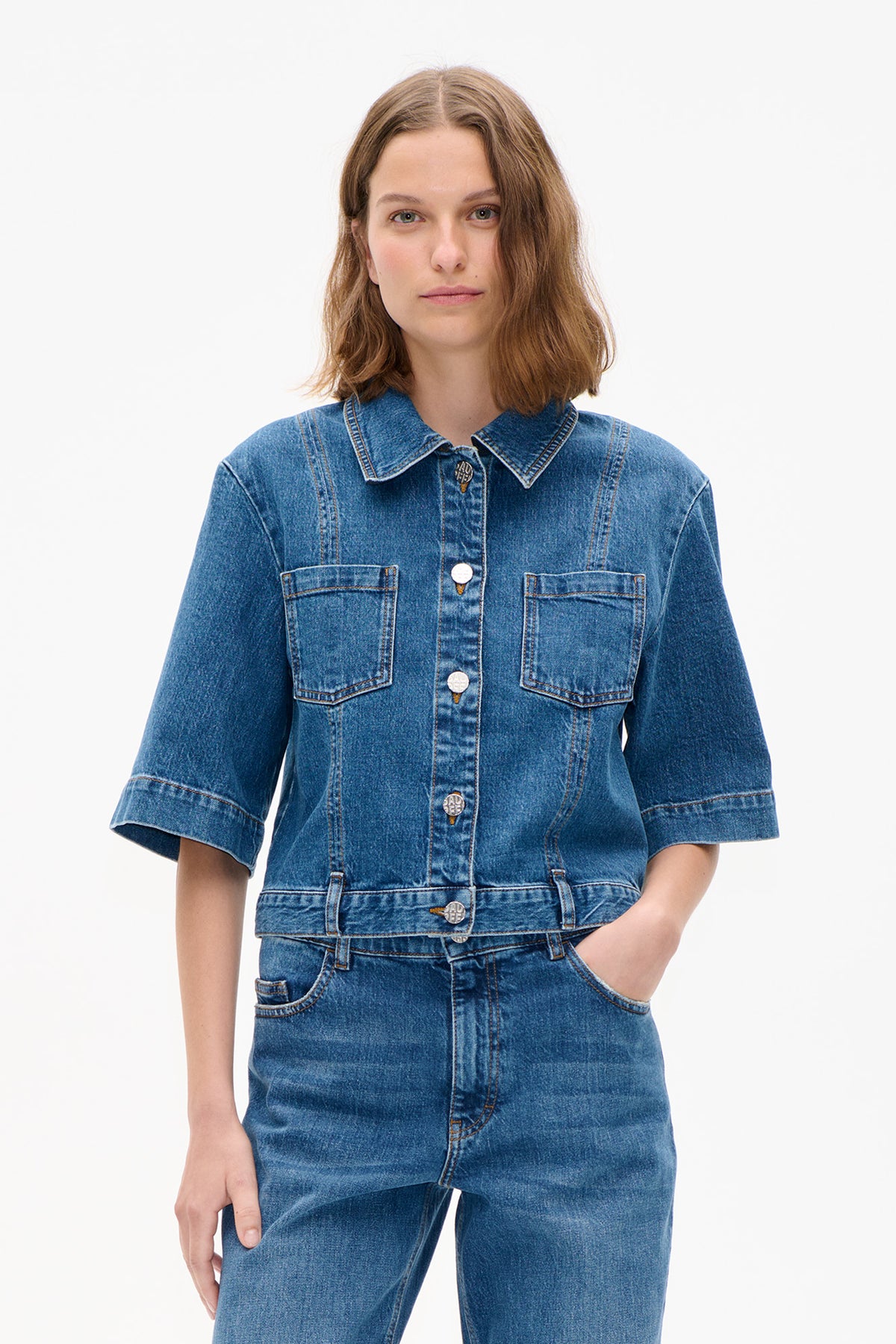 Front view of a model wearing a blue denim jacket with short sleeves, pointed collar, chest pockets and metal buttons, paired with jeans.