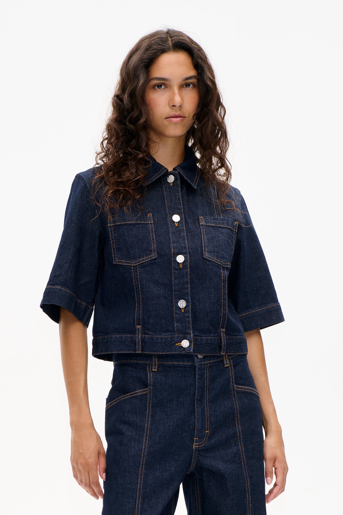 Vue de face d'une veste en denim indigo foncé à manches courtes, deux poches poitrine, boutons argentés et surpiqûres contrastantes.