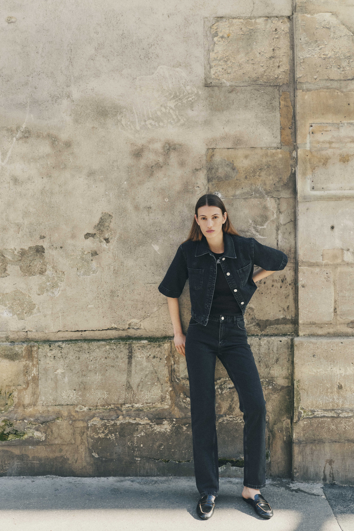 Full-length campaign portrait of a model in a dark denim short-sleeve jacket and matching jeans against a weathered stone wall.