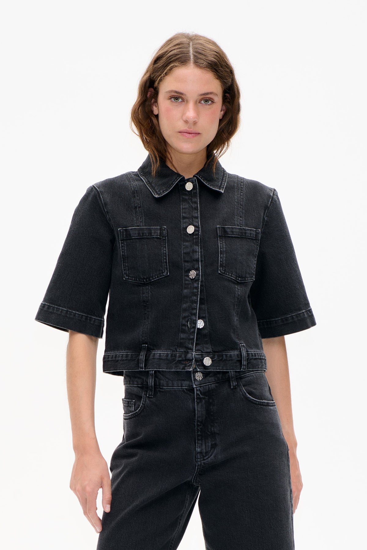 Front view of a model wearing a black washed denim jacket with short sleeves, contrasting seams, chest flap pockets and silver buttons.