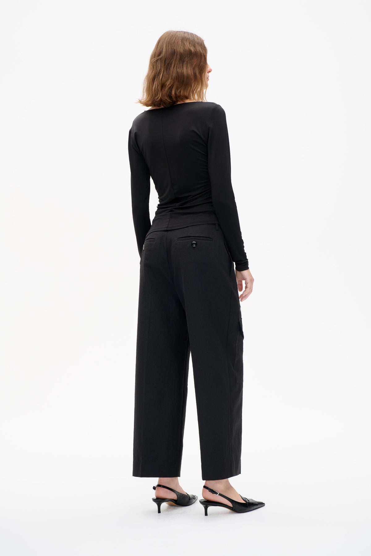 Rückansicht eines Models, das ein figurbetontes schwarzes Langarmtop und eine gekürzte, weit geschnittene Hose mit paspelierten Gesäßtaschen und Knopfverschlüssen trägt.