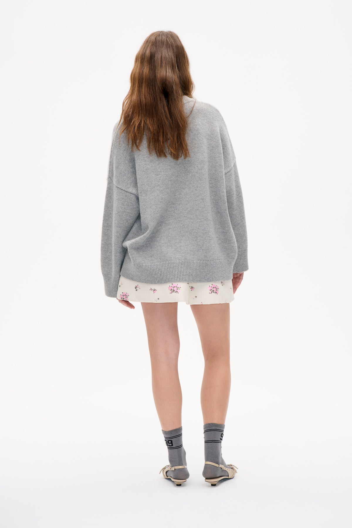 Rear view of a model wearing an oversized light grey knit sweater with a large lower-back pocket, paired with a cream floral mini skirt and striped socks.