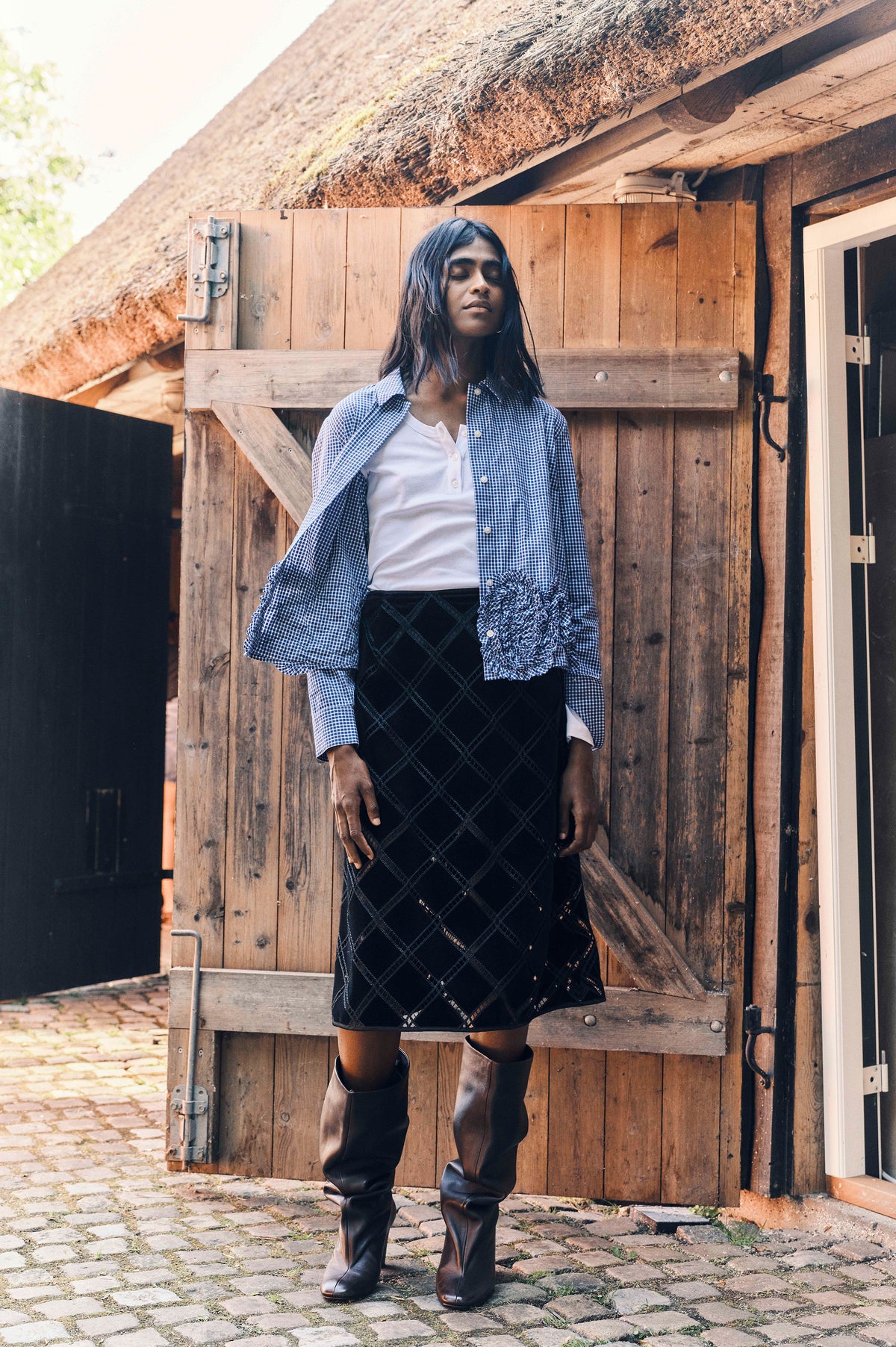Model draagt een blauw-wit geruite blouse met ruches over een wit topje, een zwarte rok met ruitmotief en hoge donkere laarzen bij een houten poort.