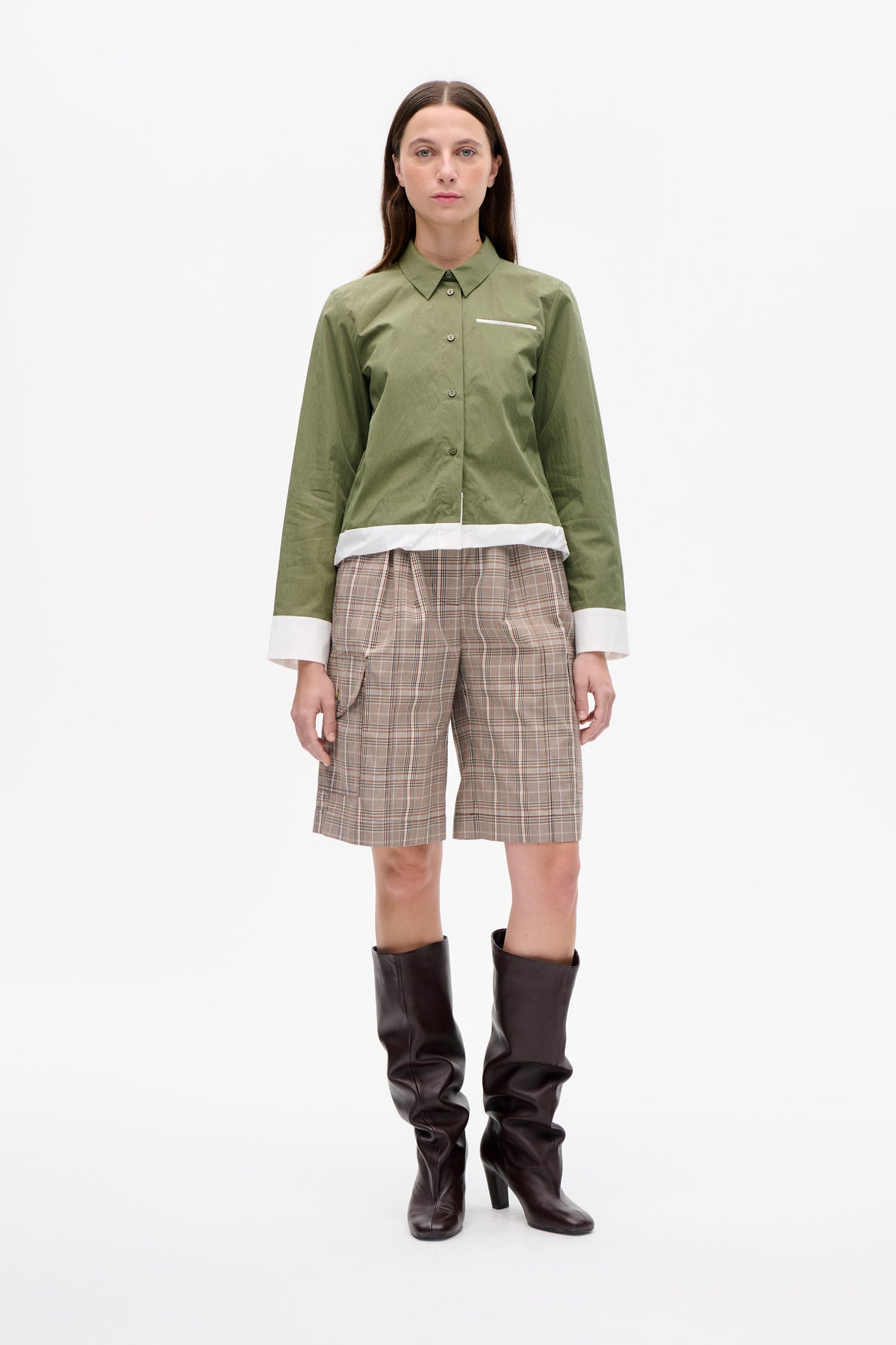 Front view of an olive green button-front shirt with white cuffs and hem, styled with beige plaid shorts and dark knee-high boots.