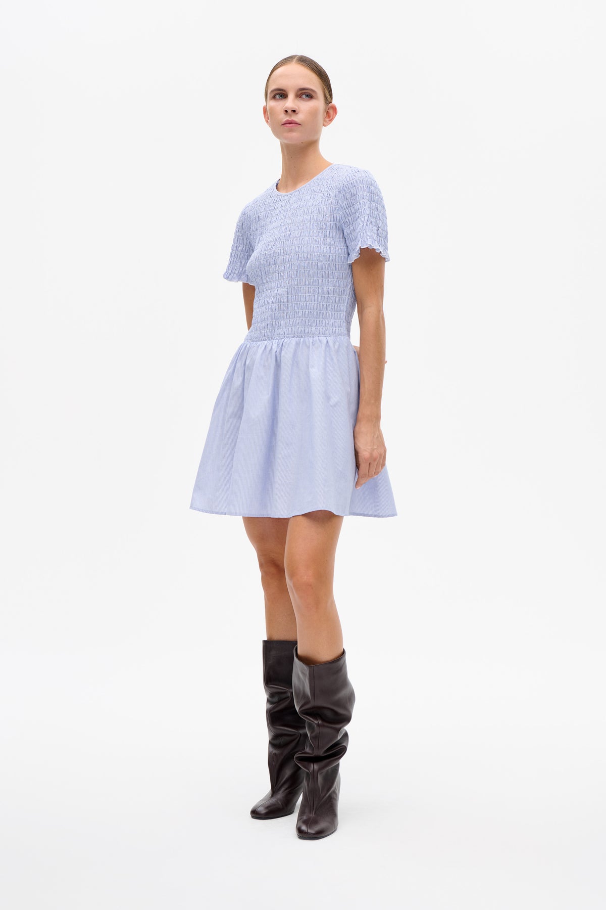 Model wearing a pale blue short-sleeved dress with a ruched bodice, ruffled sleeve edges and a subtly striped flared skirt, paired with dark knee-high boots.