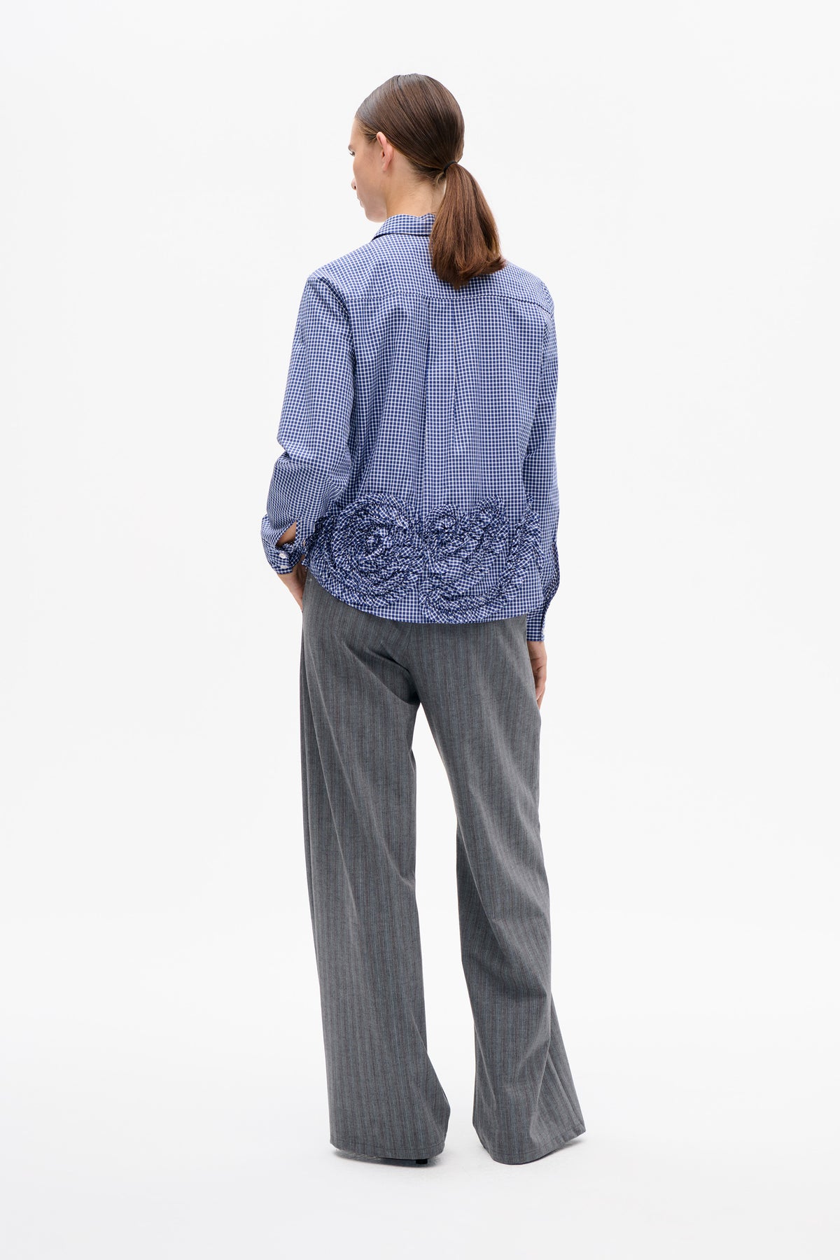 Achteraanzicht van een blauw-witte geruite blouse met lange mouwen en een ruchezoom, gedragen met een grijze broek met wijde pijpen.