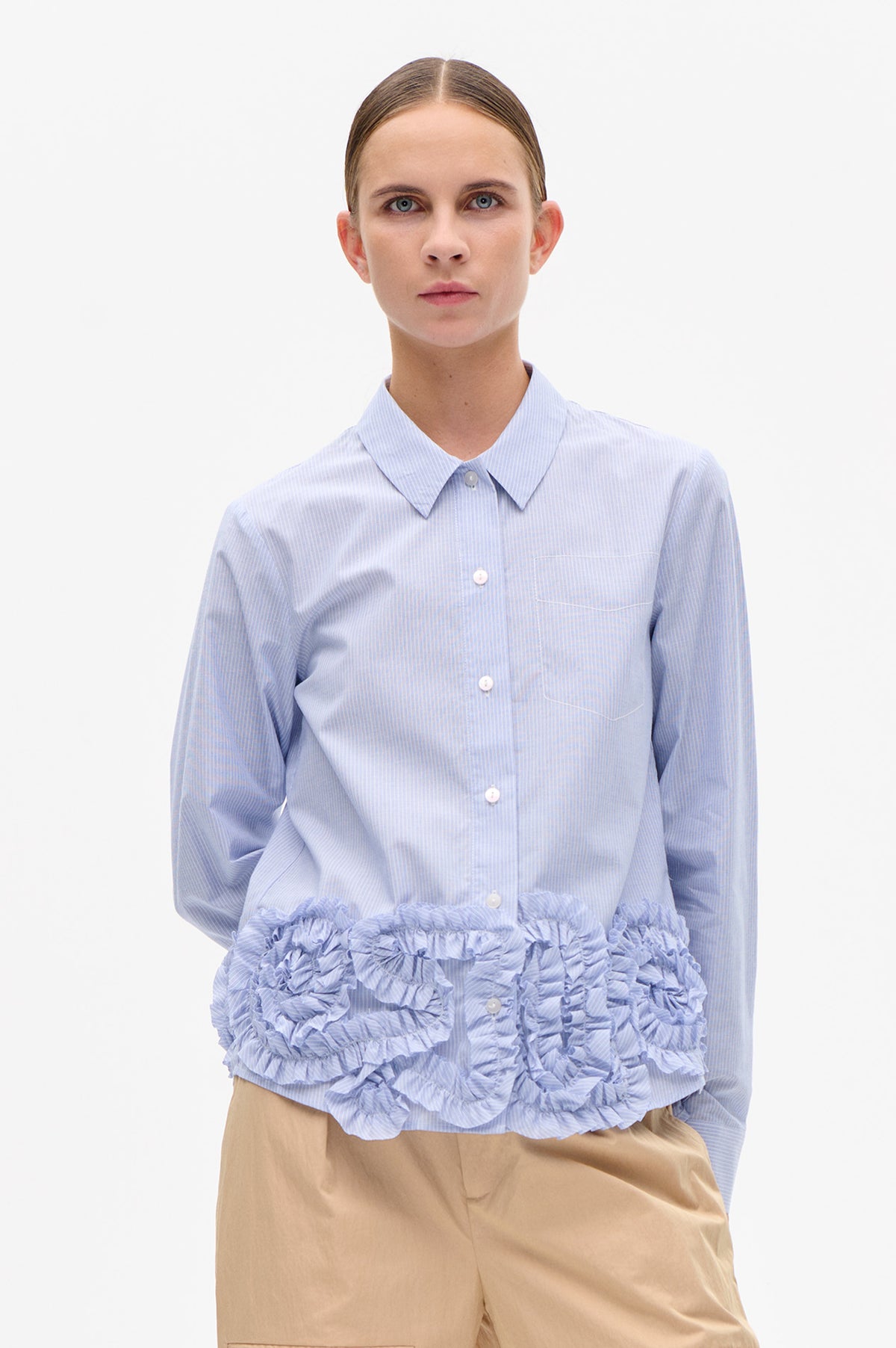 Front view of a light blue and white striped button-up shirt with a pointed collar, chest pocket and layered ruffled hem, worn with beige trousers.