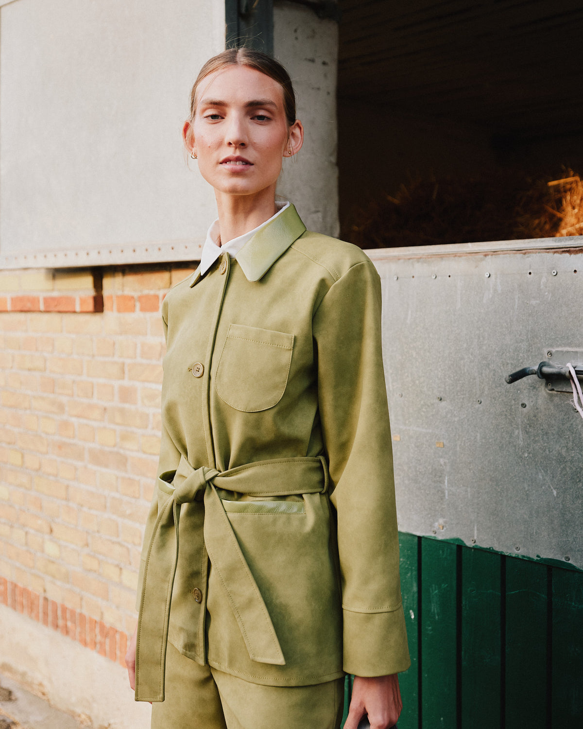 Model wearing a pale olive belted jacket with a pointed collar, button front and large patch pocket, shown from the front.