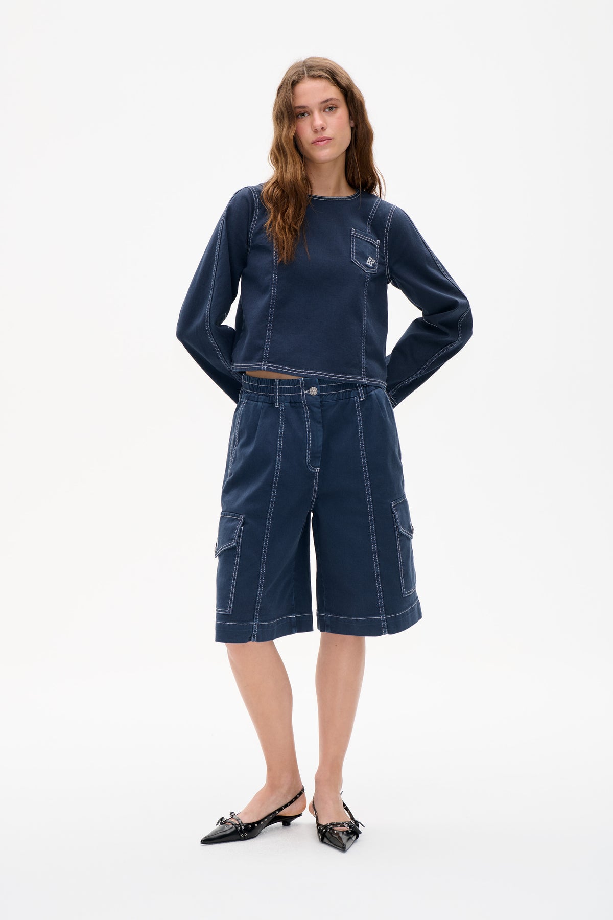 Vorderansicht eines Models, das ein marineblaues Langarmtop mit Brusttasche und dazu passende Cargo-Shorts trägt; beide mit weißen Kontrastnähten.