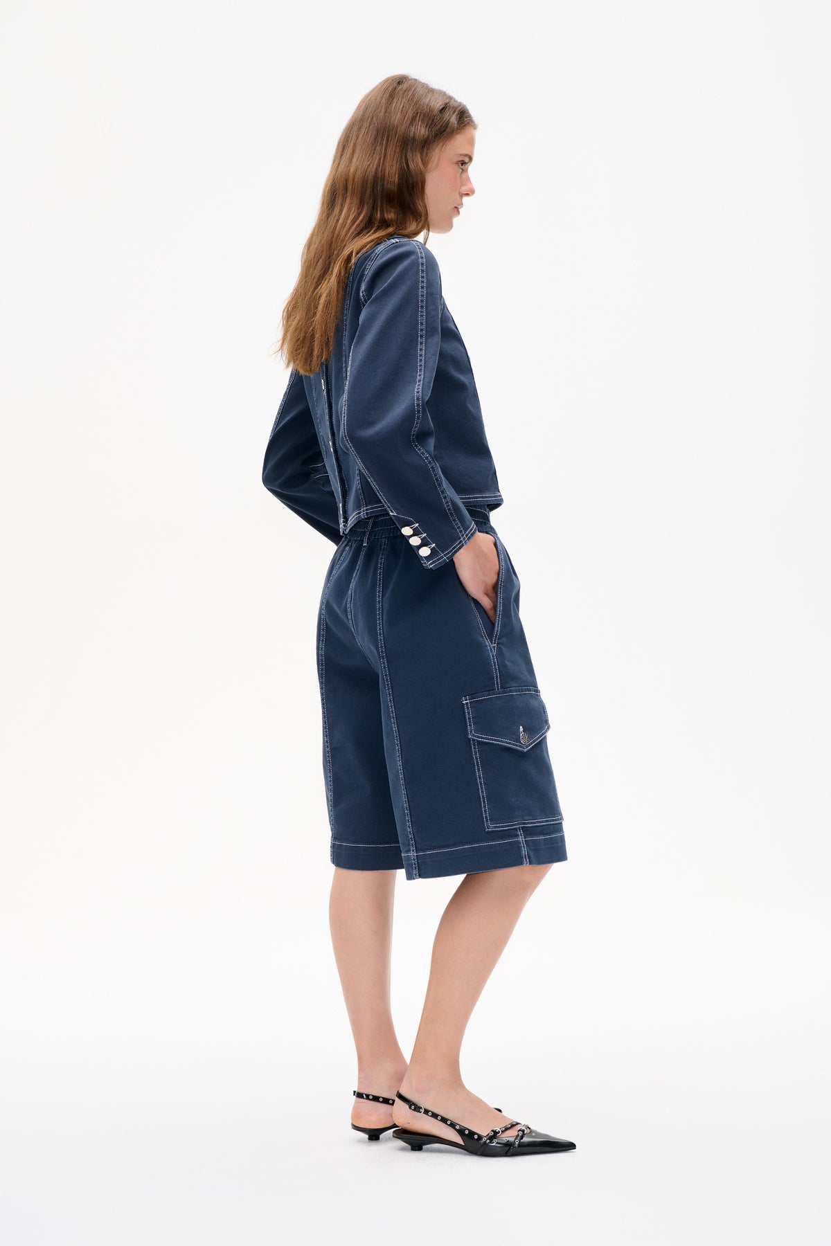 Seitenansicht eines Models, das eine marineblaue Jacke und einen passenden knielangen Rock trägt, mit Kontrastnähten, geknöpften Manschetten und einer großen aufgesetzten Tasche.