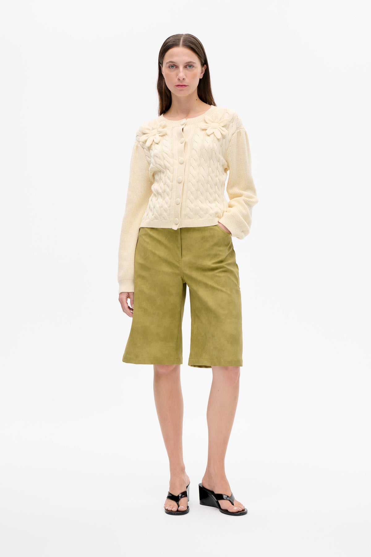 Vorderansicht eines Models, das einen elfenbeinfarbenen Zopfstrick-Cardigan mit Knöpfen und erhabenen Blumenapplikationen sowie olivgrüne, knielange Shorts trägt.