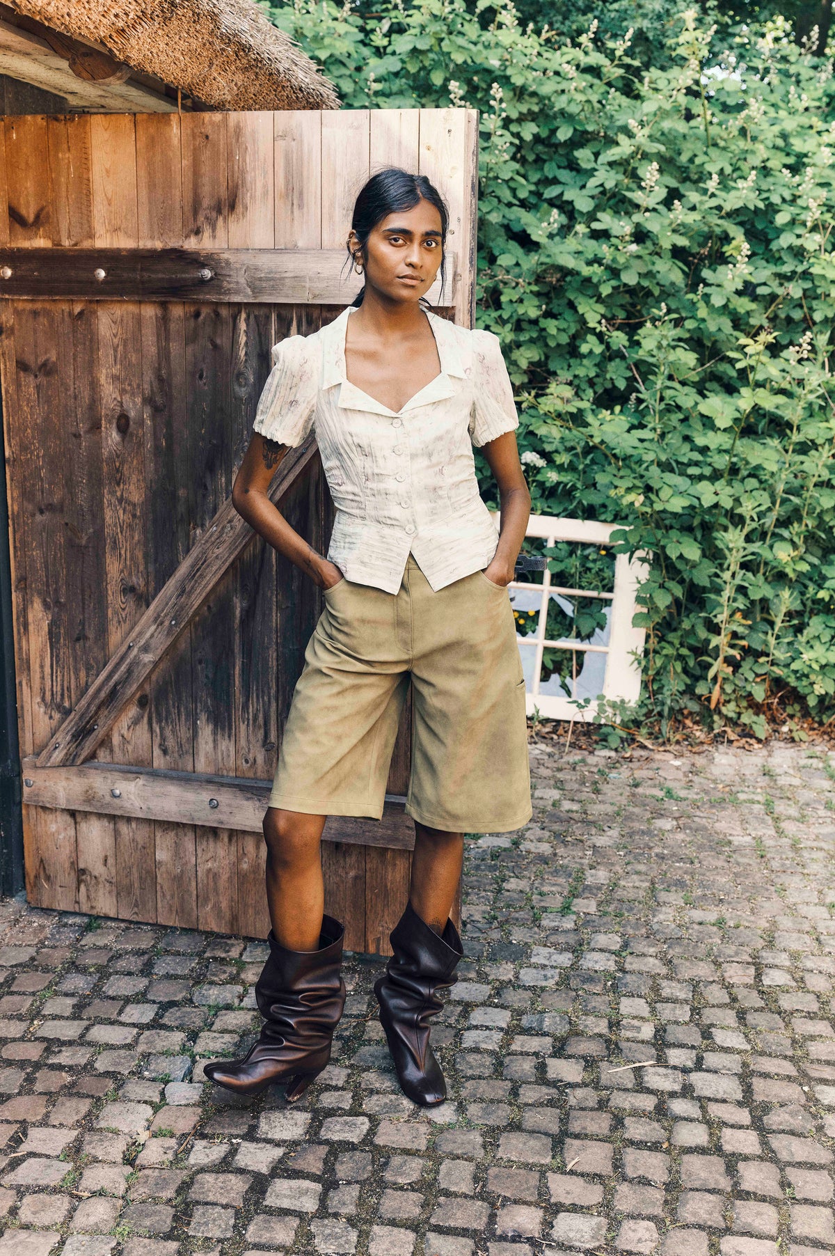Model trägt eine hell gemusterte, kurzärmelige Bluse mit durchgehender Knopfleiste, khakifarbene Shorts und dunkle, kniehohe Stiefel neben einem Holzschuppen.