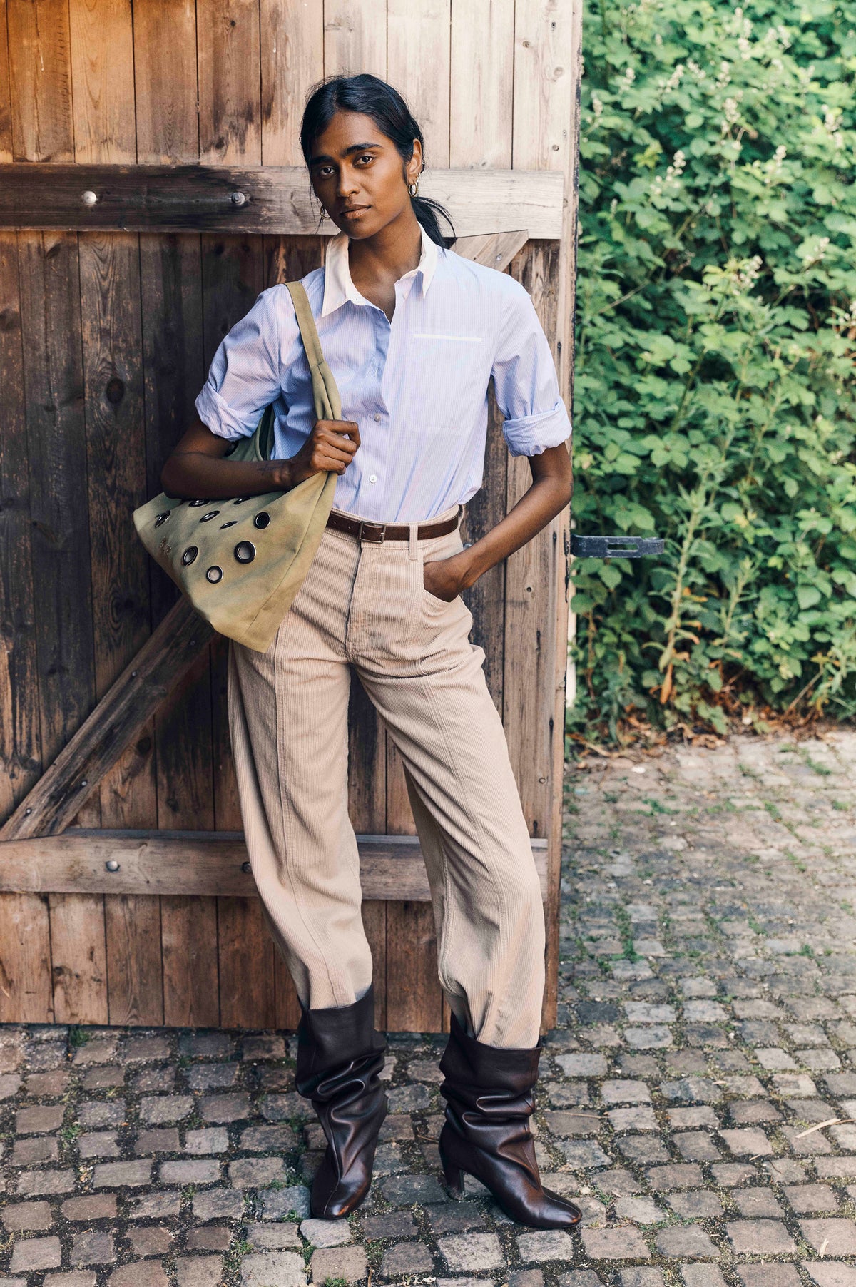 Model draagt een lichtblauwe, gestreepte blouse met knopen en een beige ribfluwelen broek, en heeft een kaki tote bij zich naast een houten muur