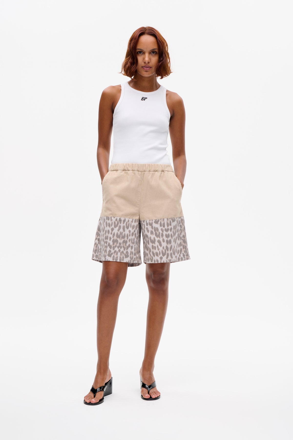 Model wearing a white ribbed tank top with a small chest logo and beige shorts with leopard-print panels.