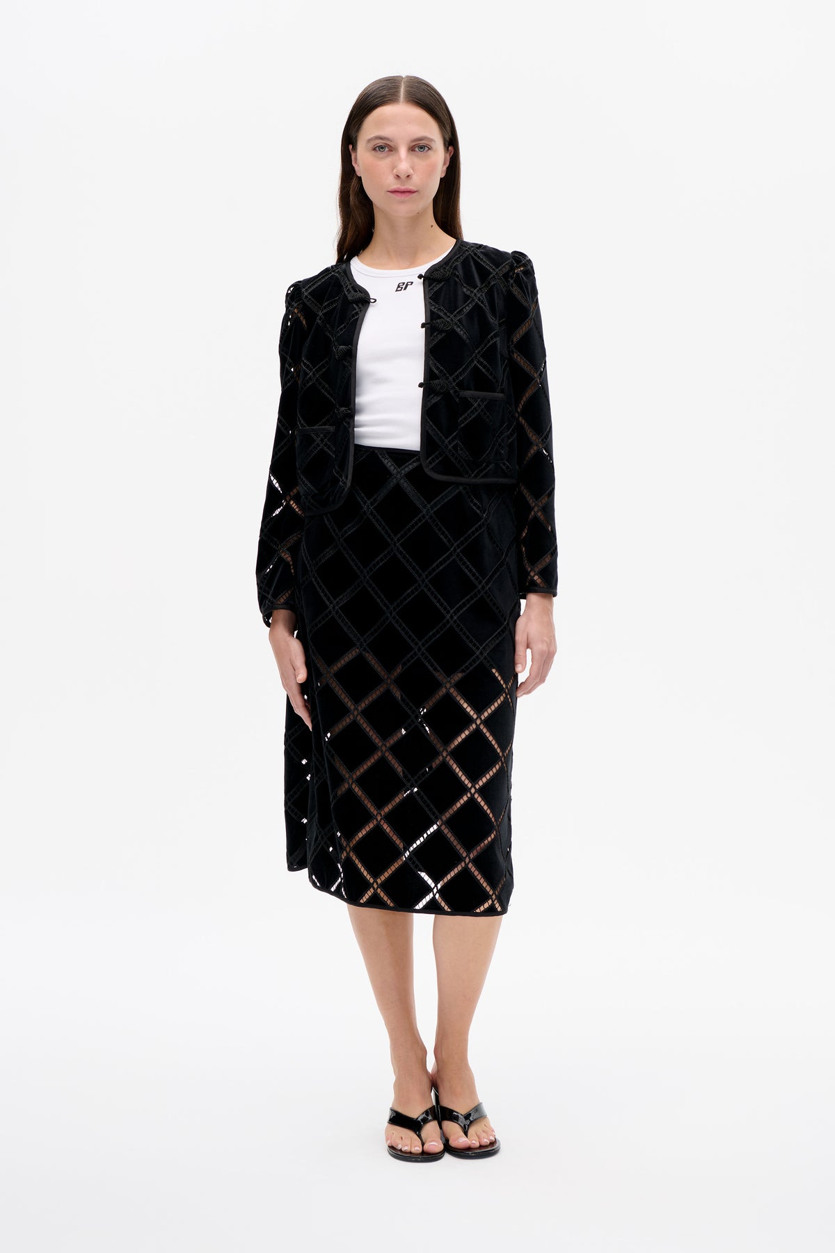 Front view of a model wearing a black knee-length jacket with a geometric openwork pattern and toggle closures over a white top.