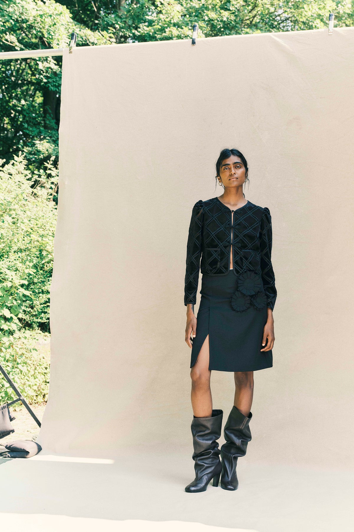 Model in a black lattice-stitched jacket, rosette-detailed skirt and tall boots against a beige backdrop with leafy greenery