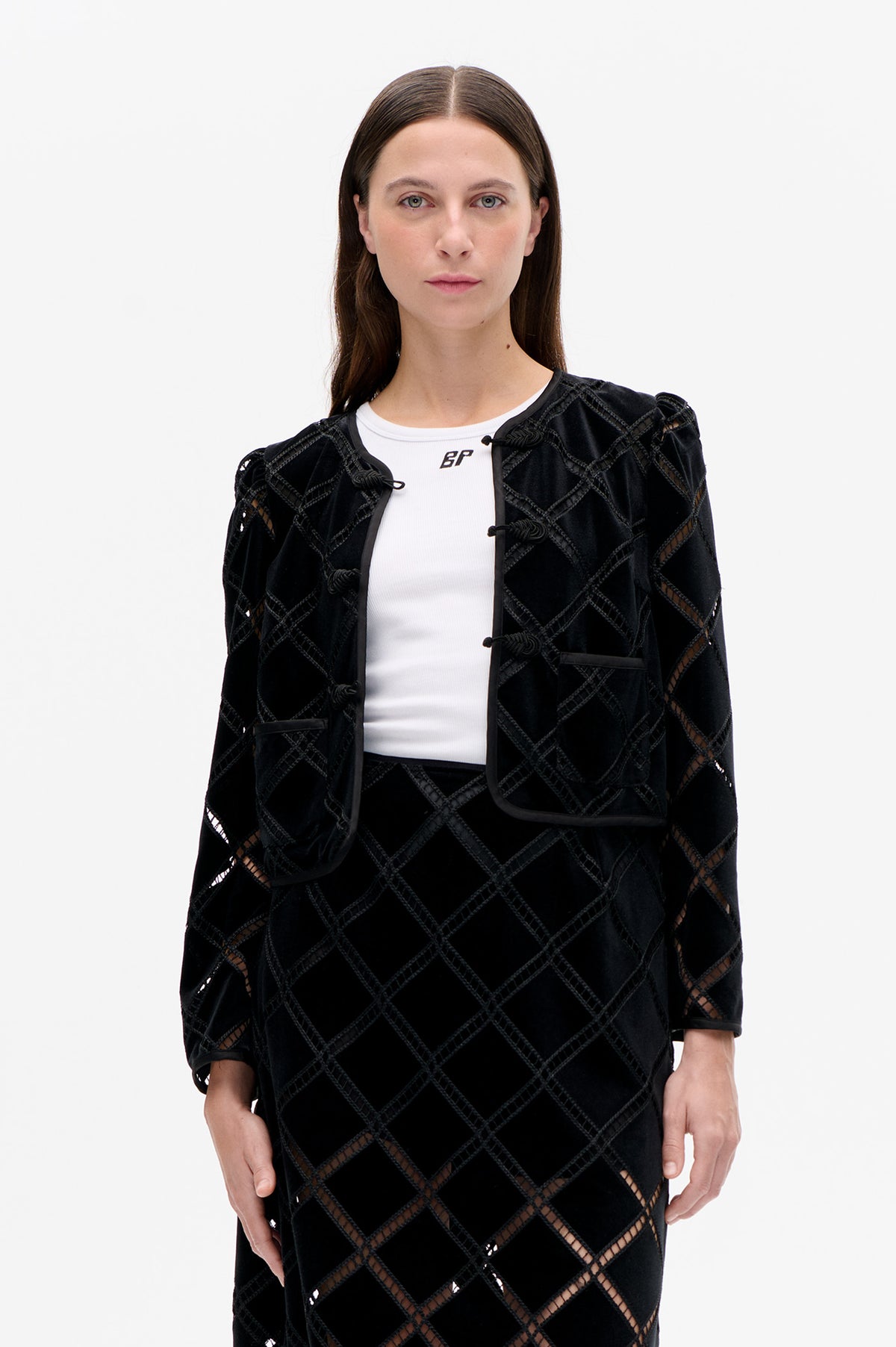 Front view of a black velvet jacket with geometric openwork lattice, toggle closures and patch pockets, worn over a white ribbed T-shirt.