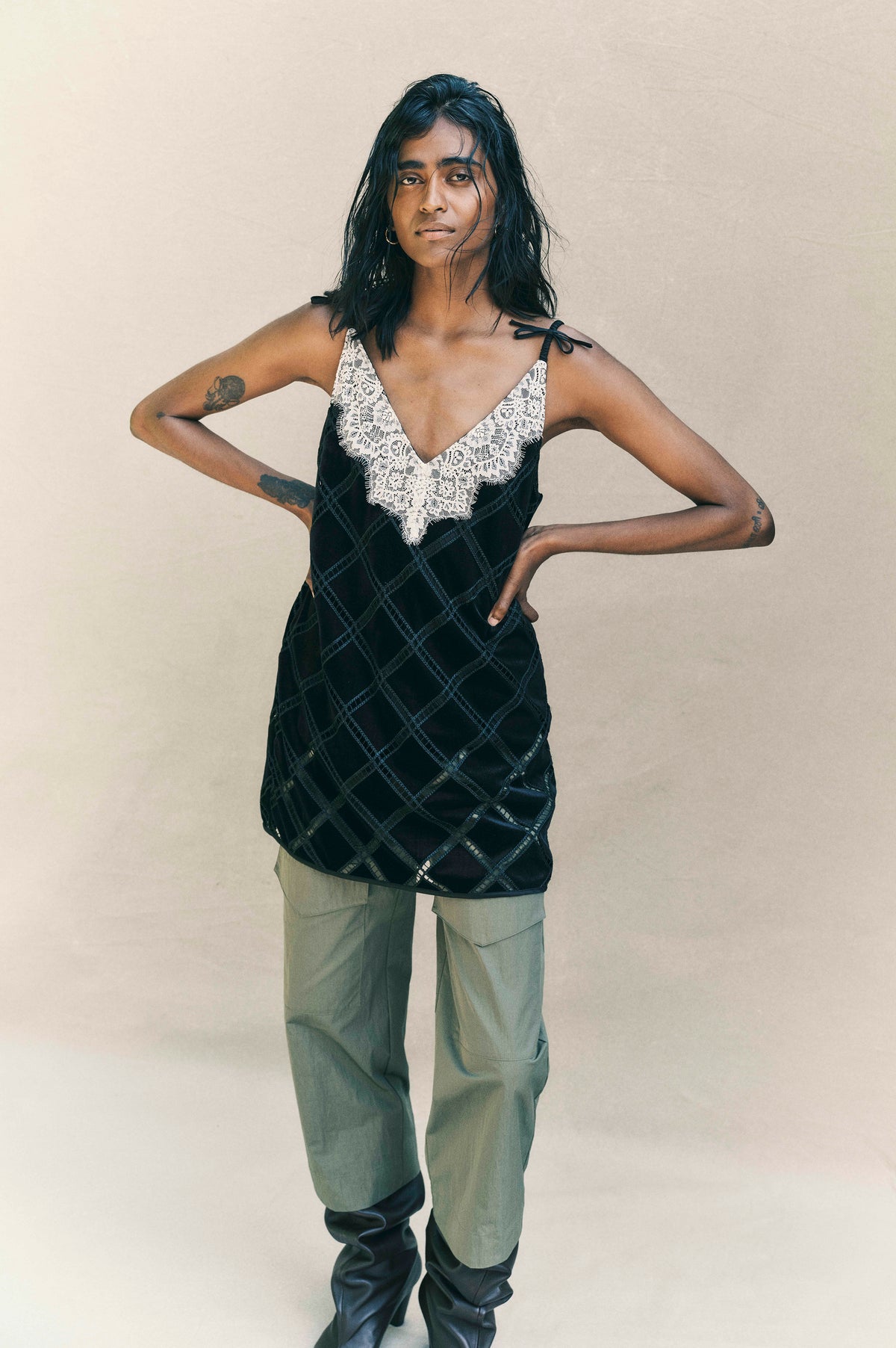 Model wearing a black sleeveless top with white lace trim and diamond stitching, paired with sage cargo trousers against a beige backdrop.