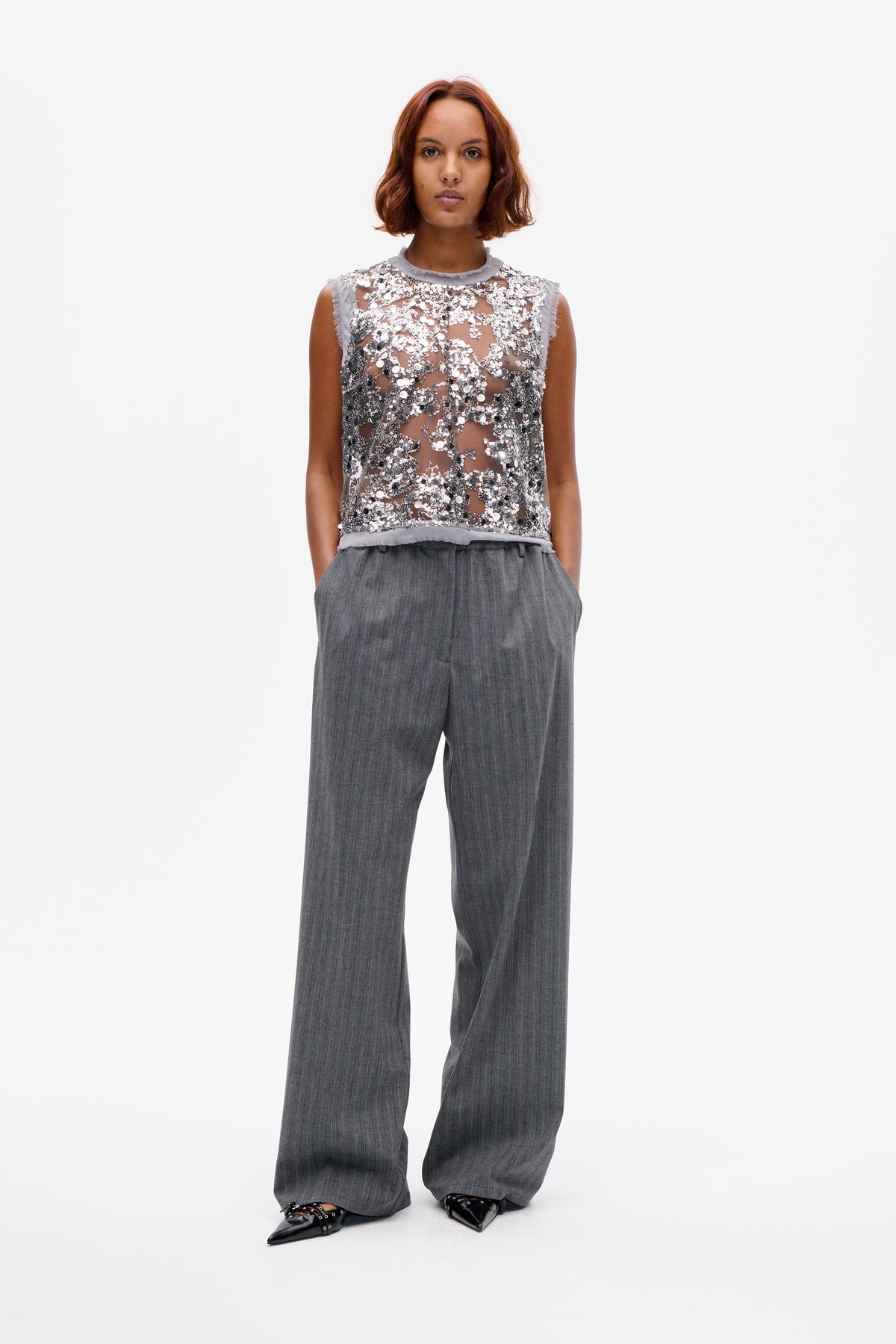 Vue de face d'un top sans manches transparent, recouvert de sequins argentés et foncés, porté avec un pantalon large gris à rayures fines.