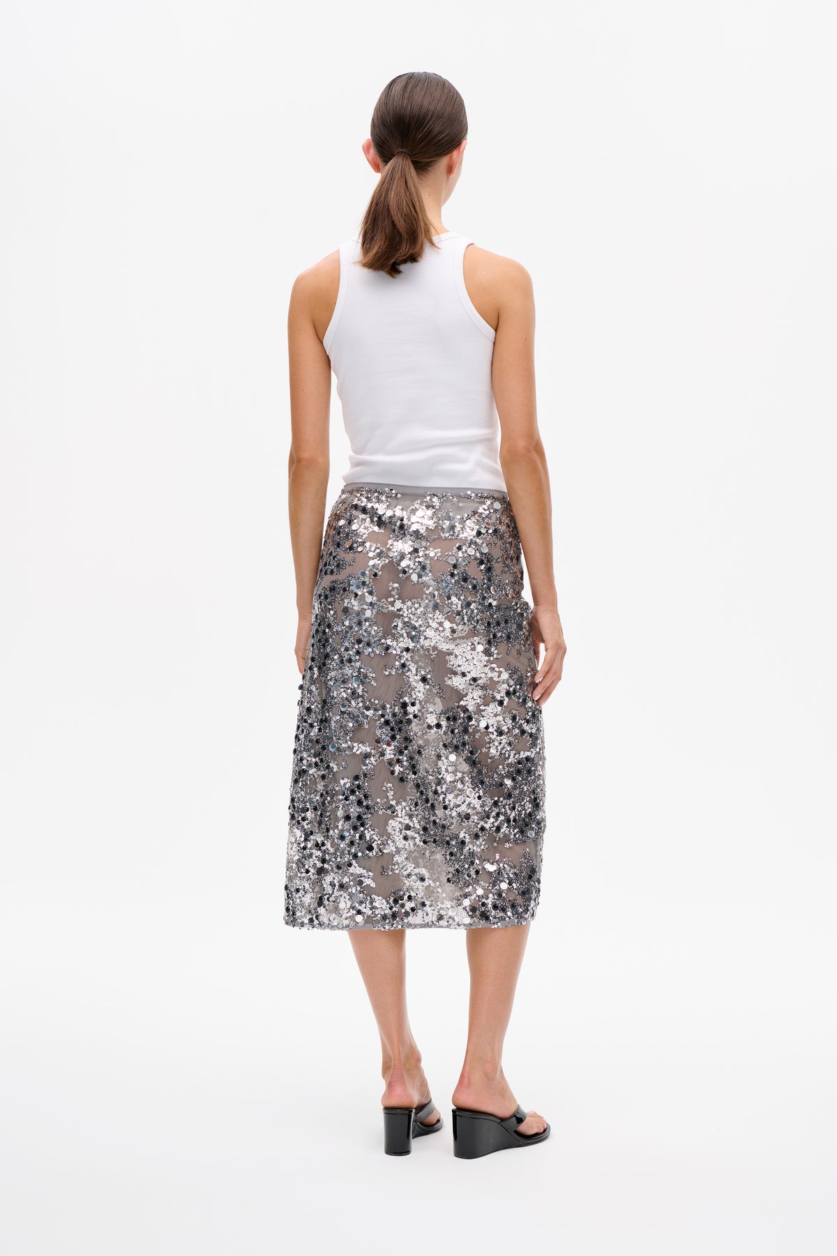 Back view of a model wearing a white ribbed sleeveless top and a knee-length sheer skirt covered in silver and black sequins.