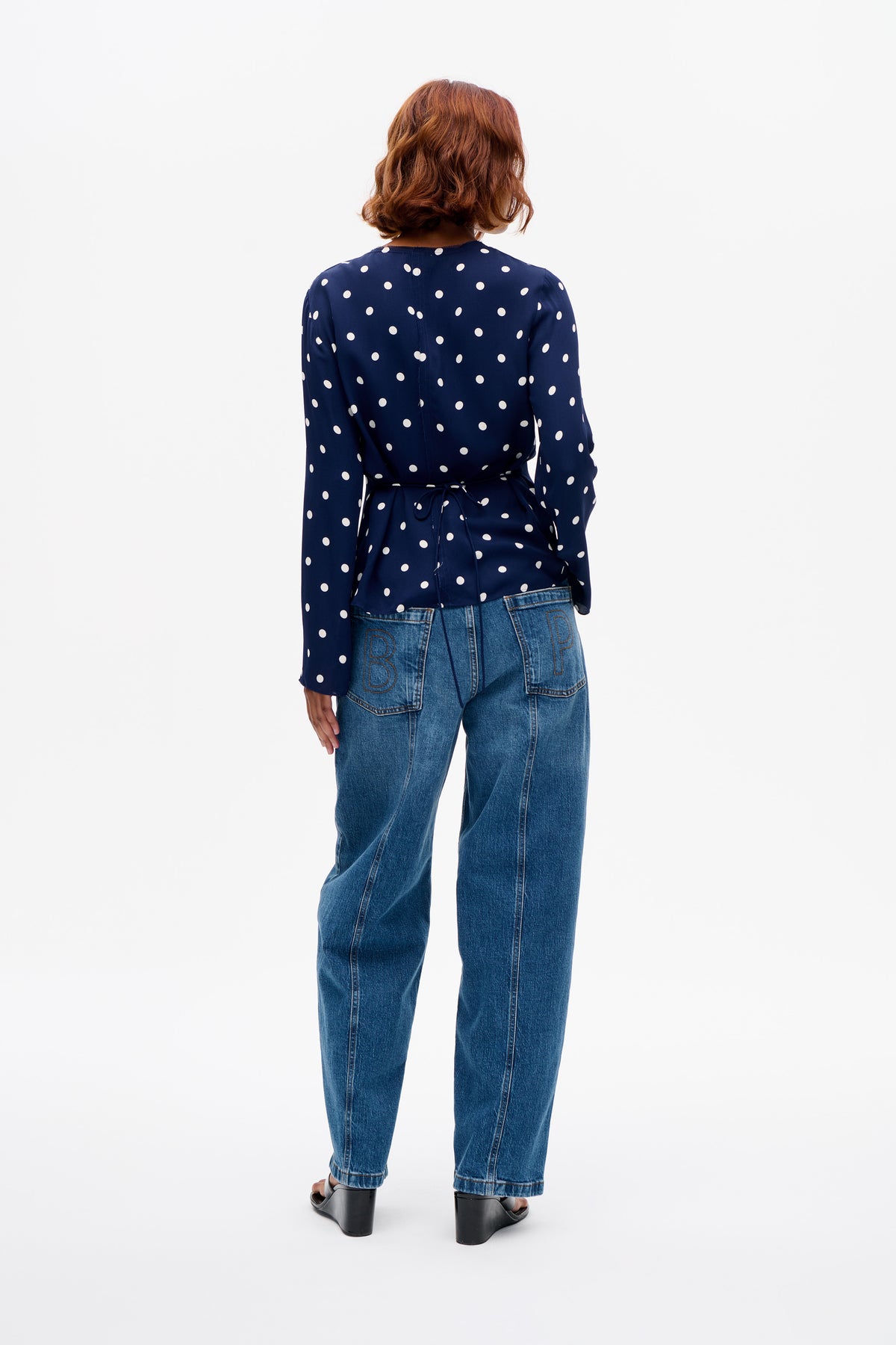 Rückansicht einer marineblauen, langärmeligen Bluse mit weißen Pünktchen und Taillengürtel, kombiniert mit blauen Jeans.