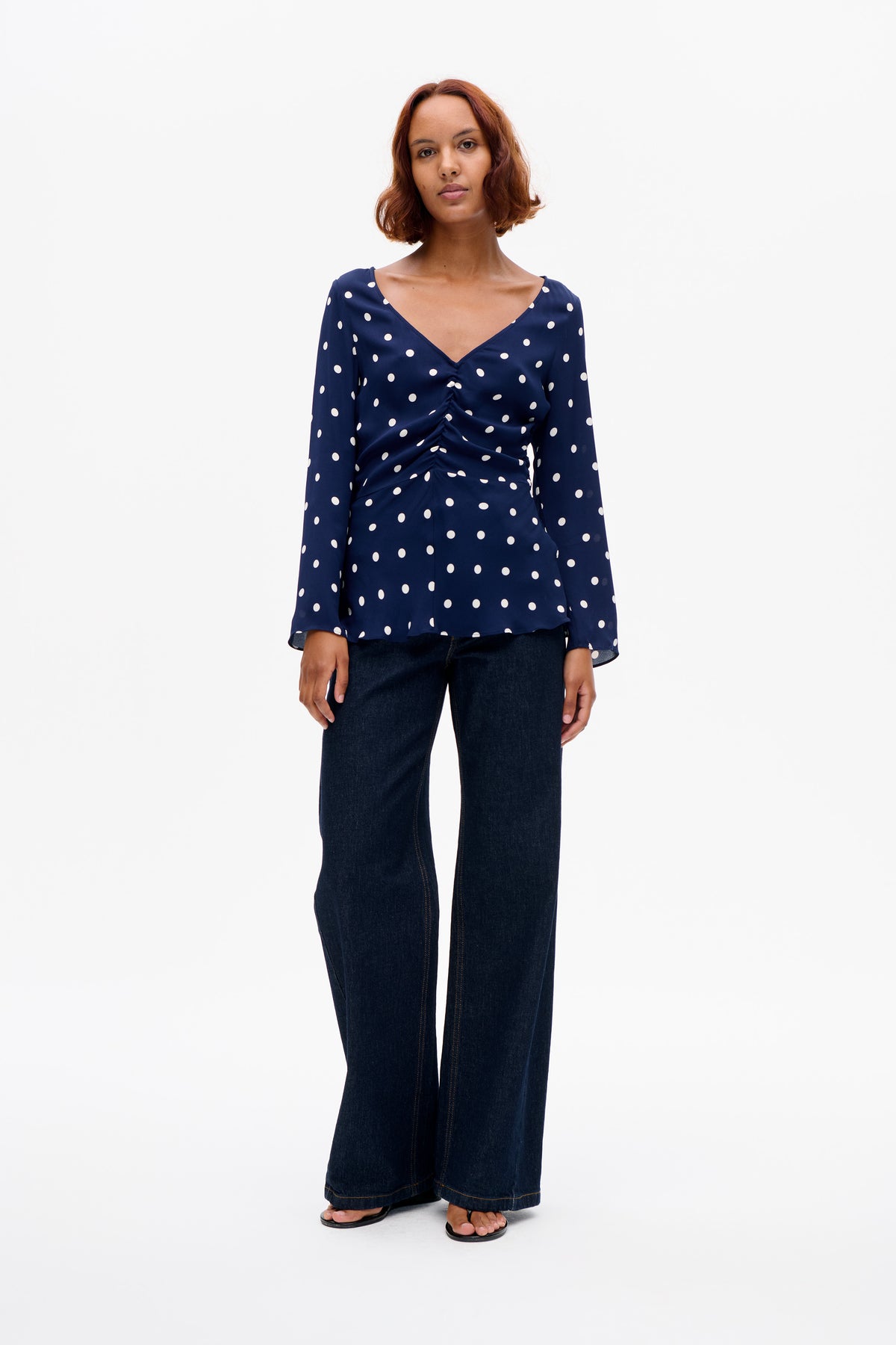 Vooraanzicht van een marineblauwe blouse met lange mouwen en witte stippen, diepe V-hals en geplooid voorpand, gecombineerd met een donkere jeans met wijde pijpen.