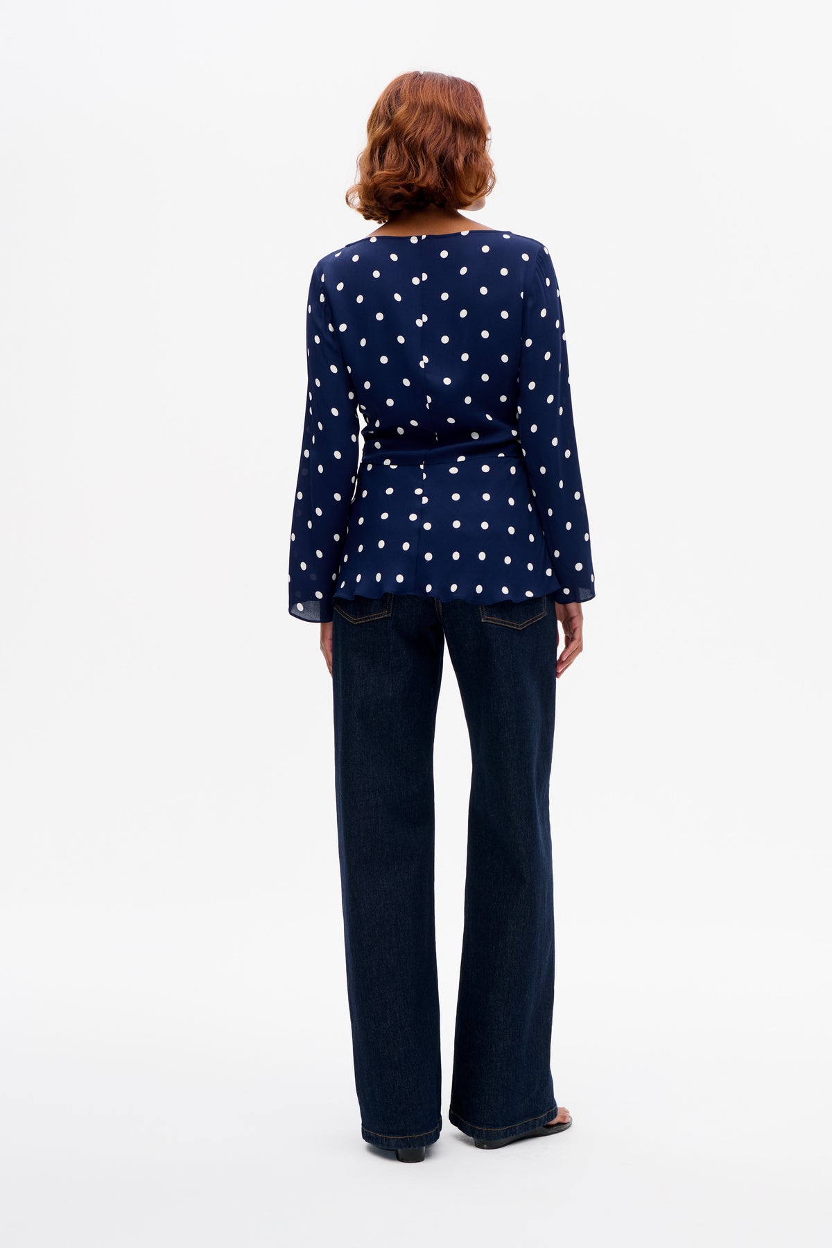 Achteraanzicht van een marineblauwe blouse met lange mouwen en witte stippen, met een uitlopende zoom, gecombineerd met donkere, wijdvallende jeans.