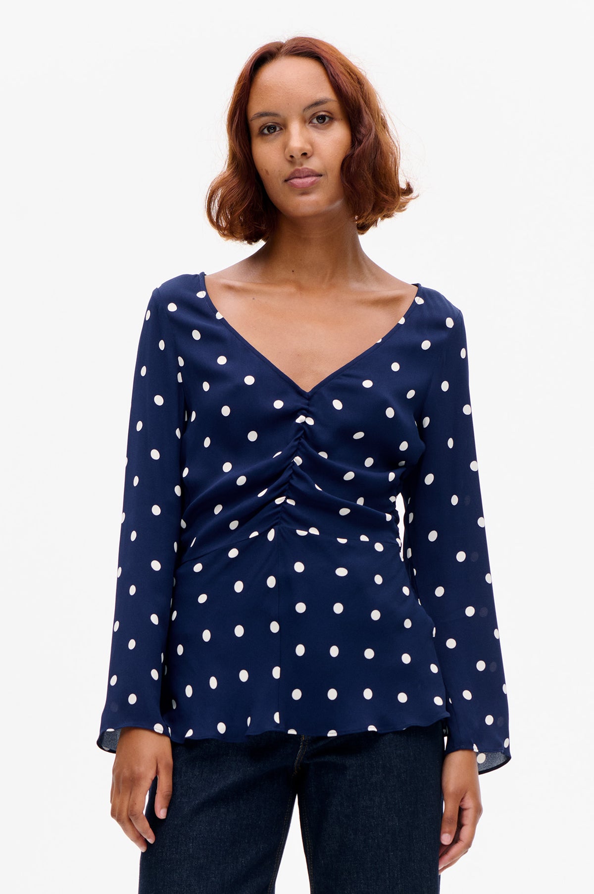 Front view of a navy blouse with white polka dots, a V-neckline and gathered detailing at the front.