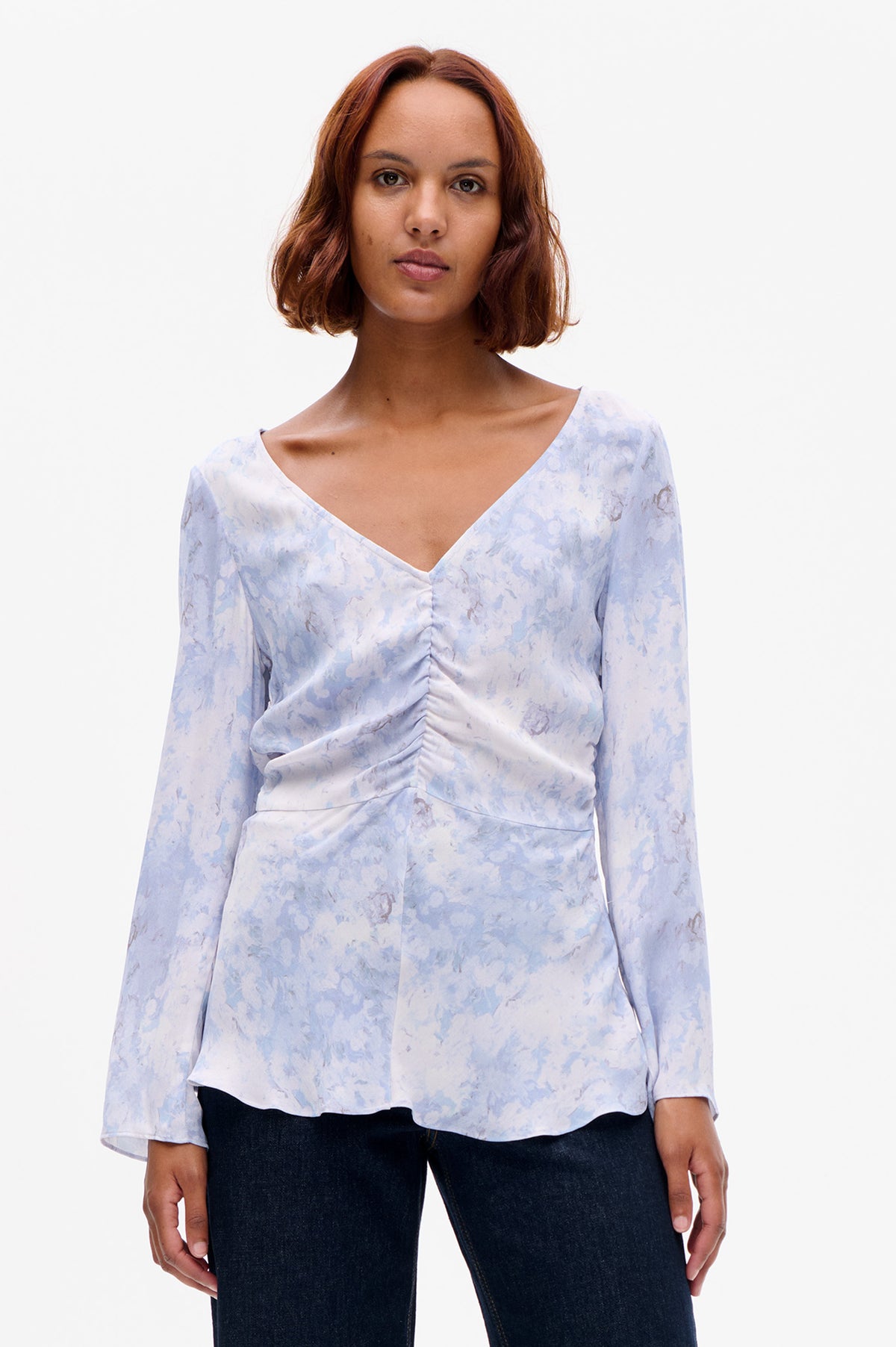 Front view of a pale blue and white abstract-print blouse with a V-neckline, long sleeves and ruched centre, worn with dark jeans.
