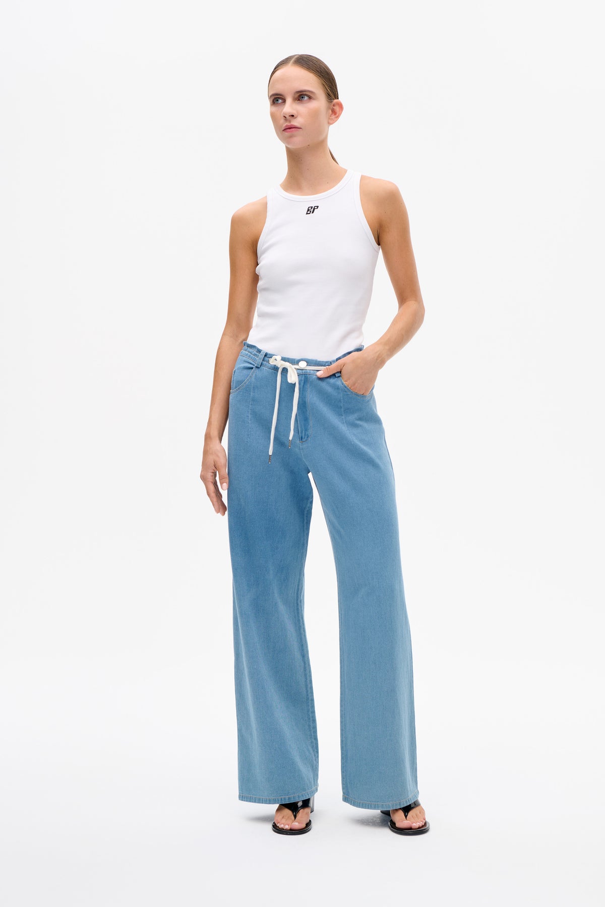 Model trägt weißes, ärmelloses, geripptes Top mit kleinem Brustlogo und hellblaue, weit geschnittene Jeans mit Kordelzugbund.