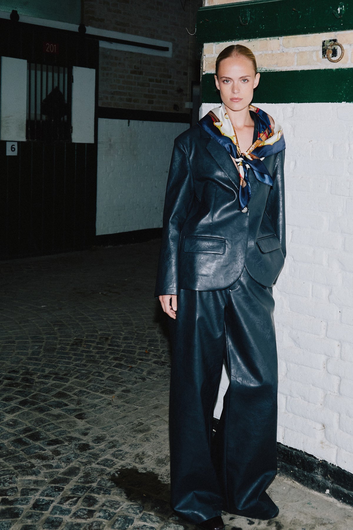 Model wearing a glossy dark navy oversized blazer and wide-leg trousers with a multicoloured patterned scarf, standing beside a brick doorway.