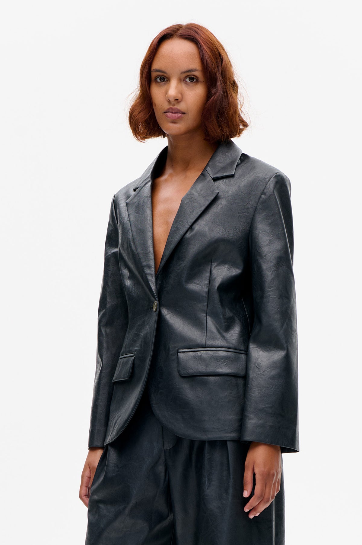 Front view of a model wearing a dark charcoal blazer with notched lapels, a single button, flap pockets and a softly creased sheen.