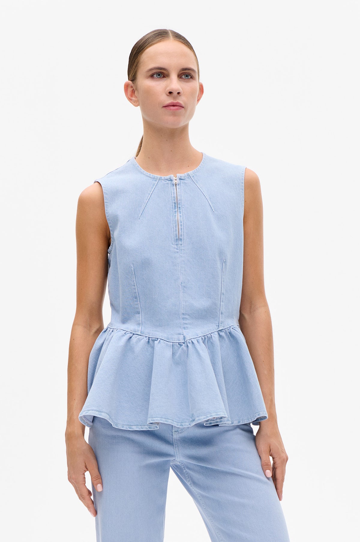 Front detail of a light blue denim sleeveless top with a front zipper, panel seams and gathered peplum hem, worn with matching jeans.