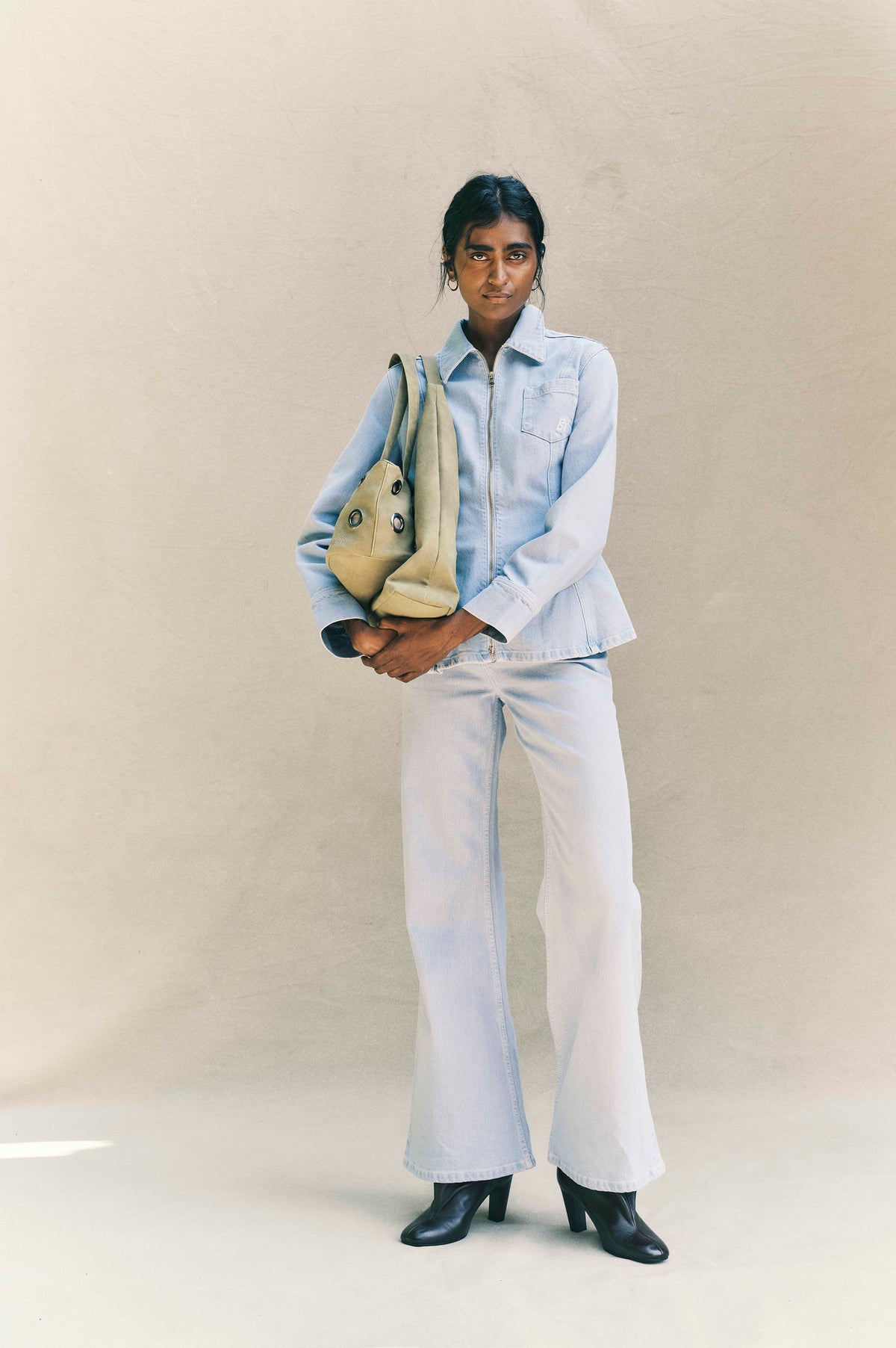 Campaign portrait of a model in a pale blue zip-front denim jacket and wide-leg jeans, carrying a tan tote and wearing black boots.