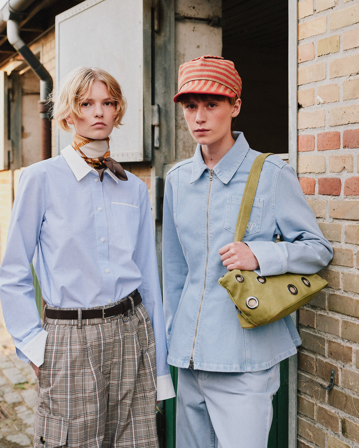 Two models pose by a brick doorway in pale blue shirts, plaid shorts, a striped cap and a light blue zip jacket with an olive bag.